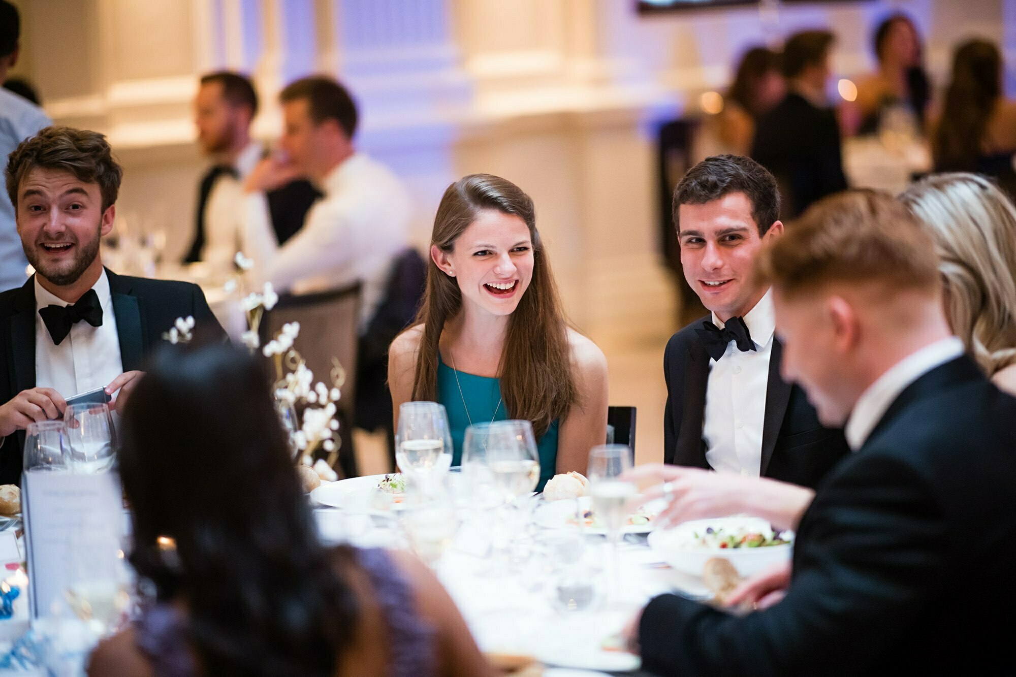 A group of people in formal attire sit around a table at a banquet. They are smiling and engaging in conversation. The table is elegantly set with glasses, plates of food, and decorative centerpieces. The background shows more guests and soft lighting. James Gifford-Mead Photography - Event Photographer London