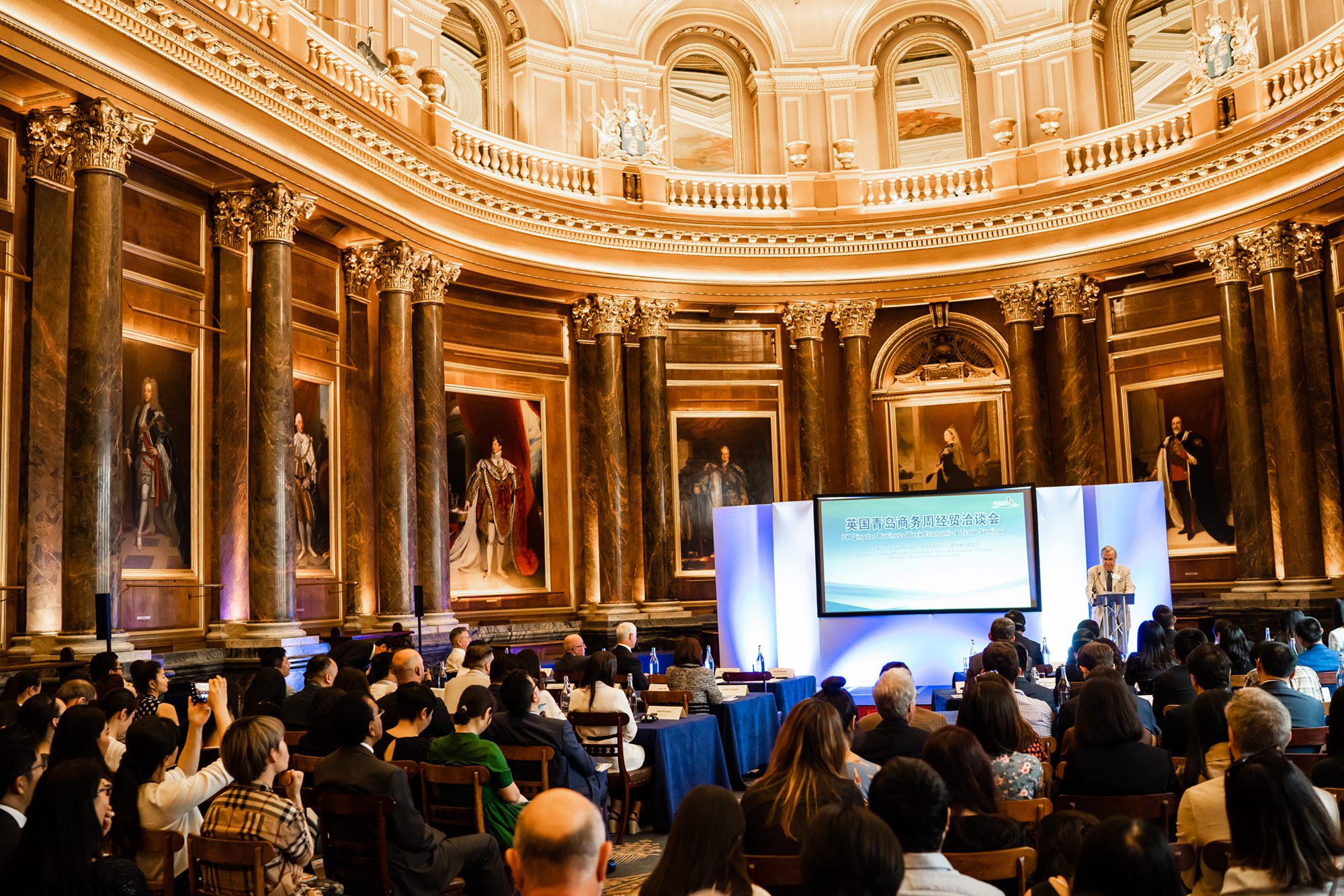 A large group of people seated in an ornate hall with marble columns and framed portraits on the walls. A speaker stands at a podium next to a large screen displaying text in Chinese. The ceiling is dome-shaped with intricate moldings. James Gifford-Mead Photography - Event Photographer London