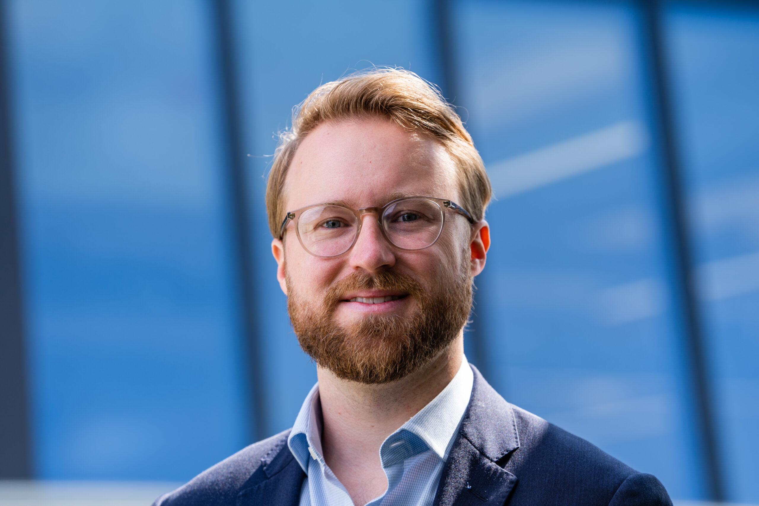 A man with a beard and glasses is smiling, dressed in a blue shirt and jacket. He stands in front of a window with a modern glass building blue facade in the background. James Gifford-Mead Photography - Event Photographer London