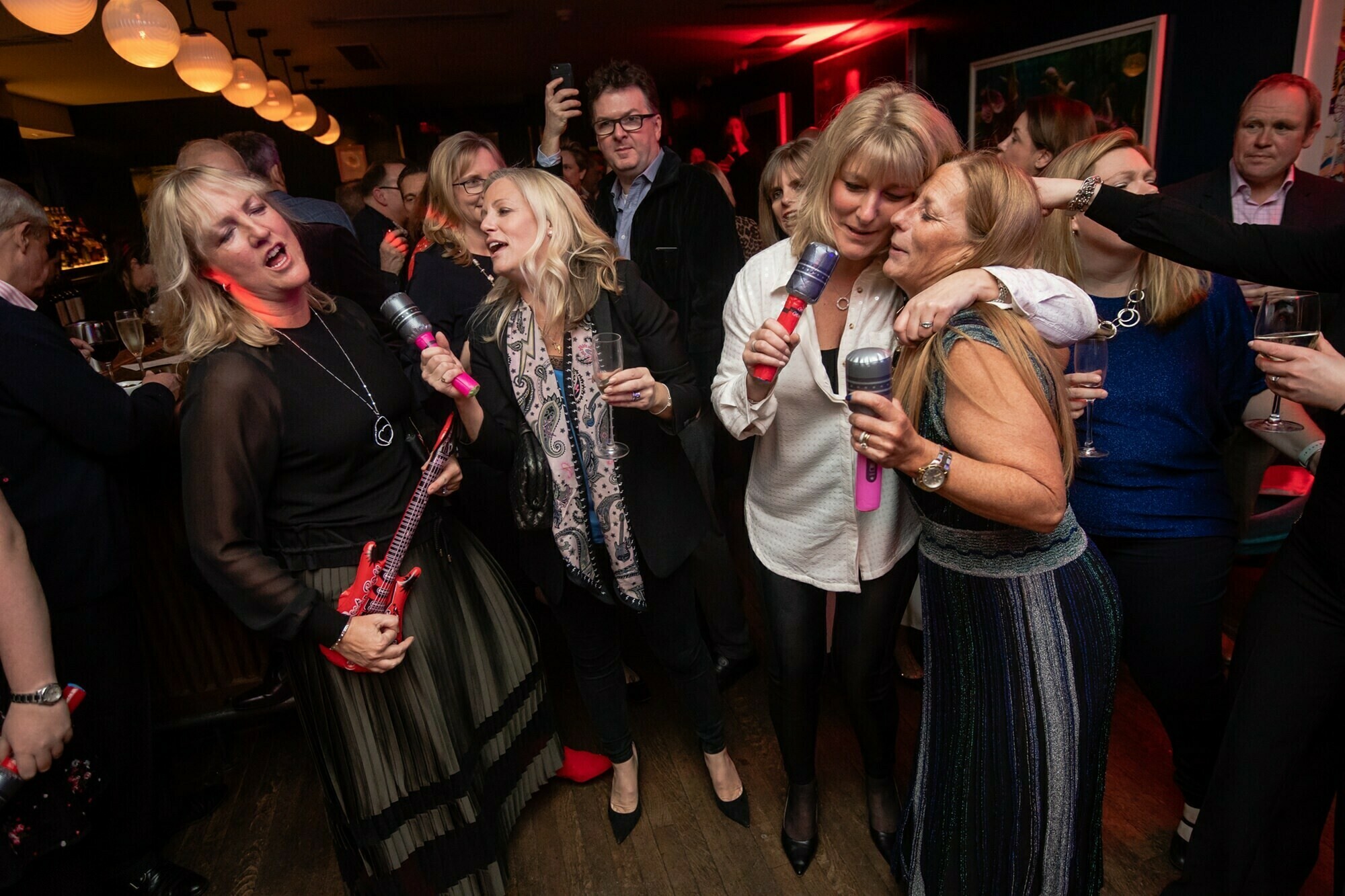 A group of people at a lively indoor party are holding inflatable microphones and guitars, singing and enjoying themselves. A woman in a long skirt sings with enthusiasm, while others gather closely, smiling and embracing. James Gifford-Mead Photography - Event Photographer London
