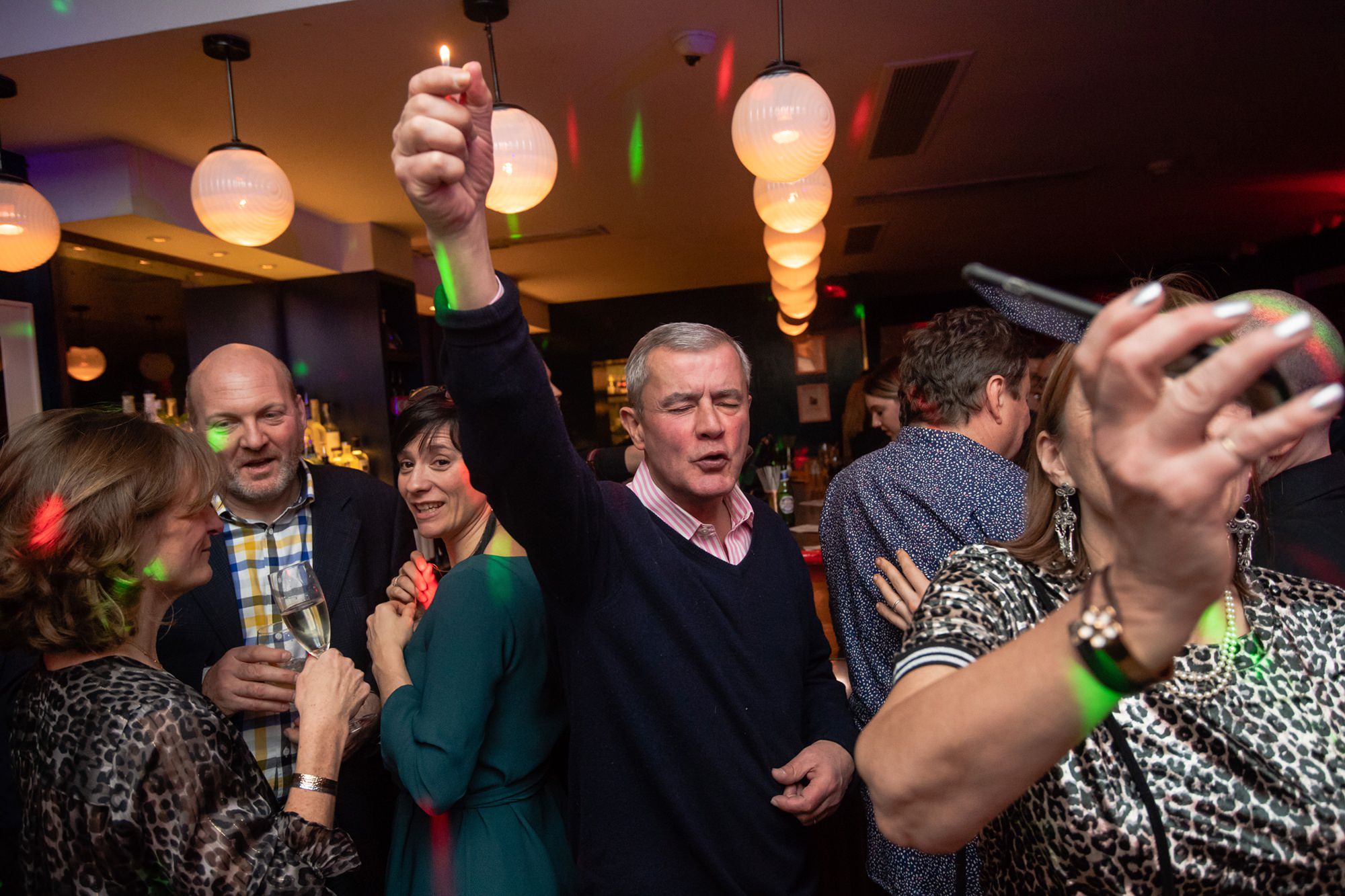 A group of people are enjoying a lively party in a dimly lit bar. A man in the center raises his fist while others around him chat and smile. The room is decorated with round lights, and a woman in the foreground takes a selfie. James Gifford-Mead Photography - Event Photographer London