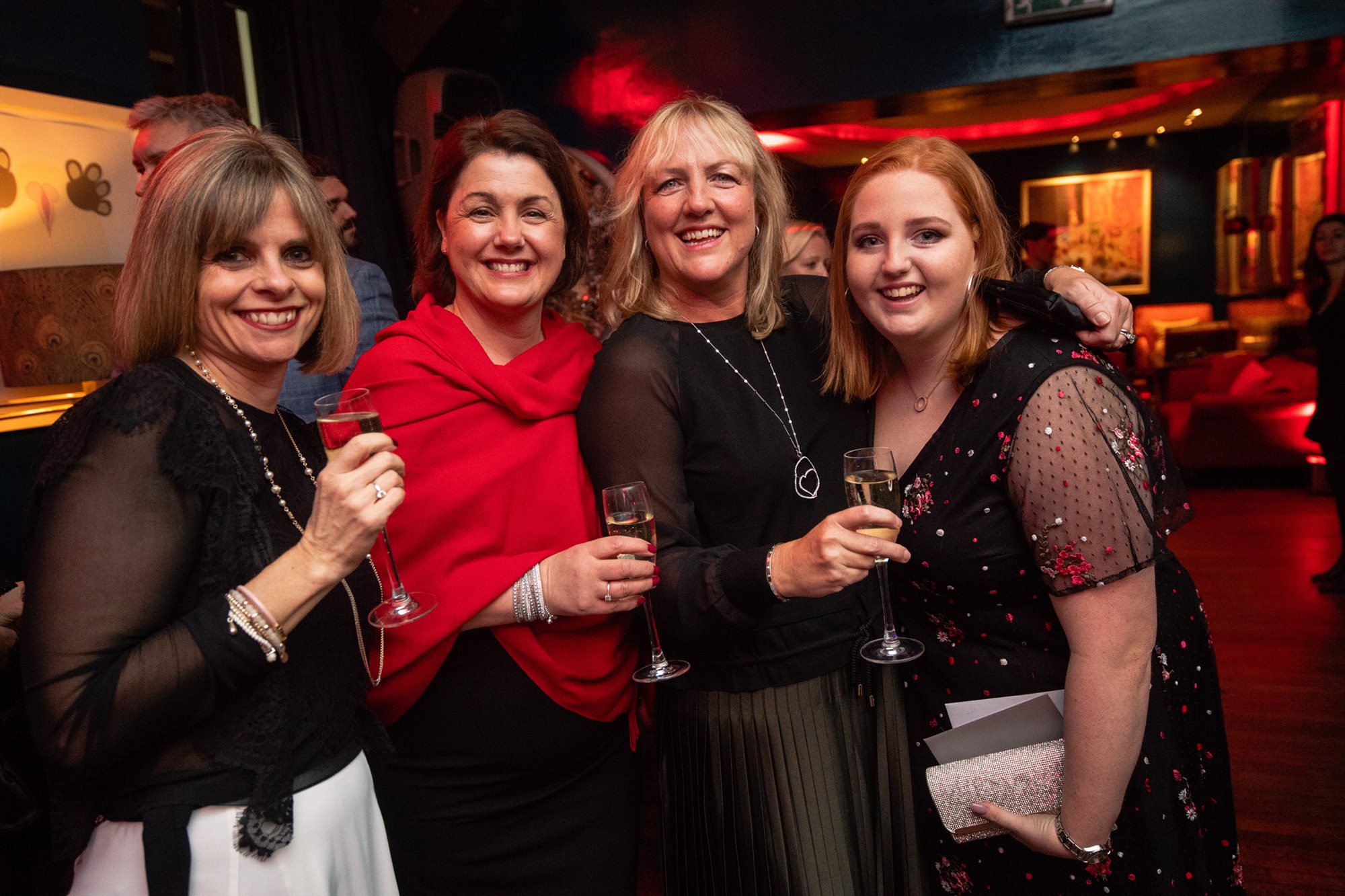 Four people are standing together at a social event, smiling and holding glasses of champagne. They are dressed in semi-formal to formal attire, with a warm, ambient setting in the background featuring dim lighting and decorative elements. James Gifford-Mead Photography - Event Photographer London