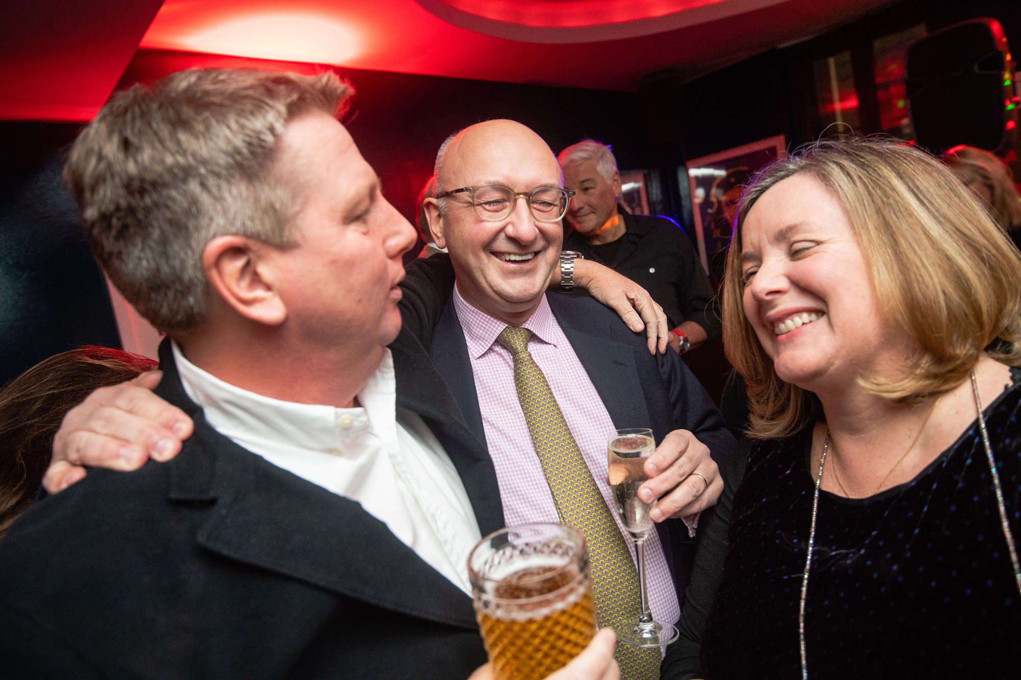 Three people, two men and a woman, are laughing and holding drinks at a social event. They appear to be in a lively atmosphere with red lighting in the background, suggesting a party or celebration. James Gifford-Mead Photography - Event Photographer London