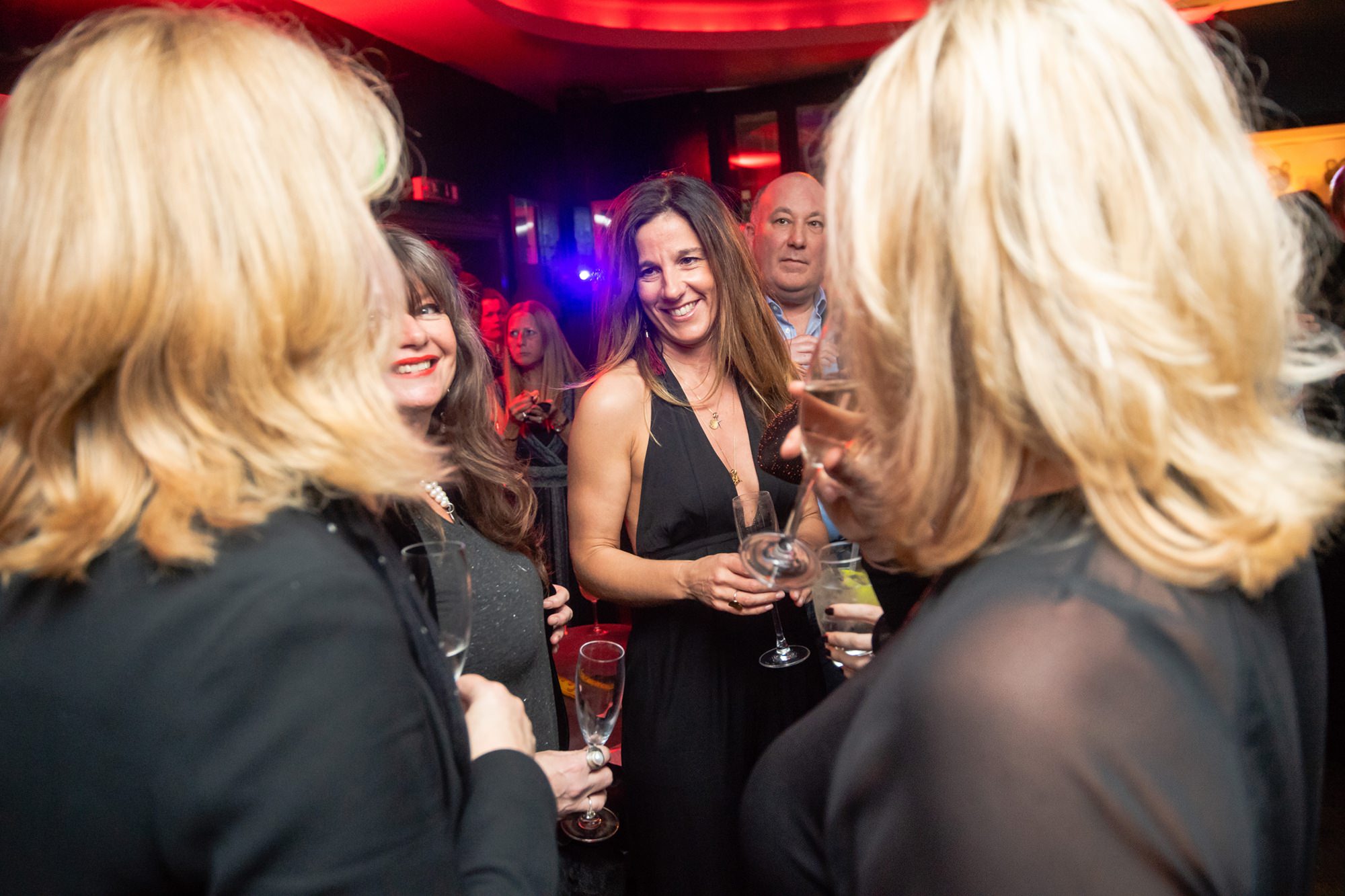 A group of people at a party, holding drinks and engaging in conversation. The setting is lively with colorful lights and several individuals smiling and dressed in evening wear. James Gifford-Mead Photography - Event Photographer London