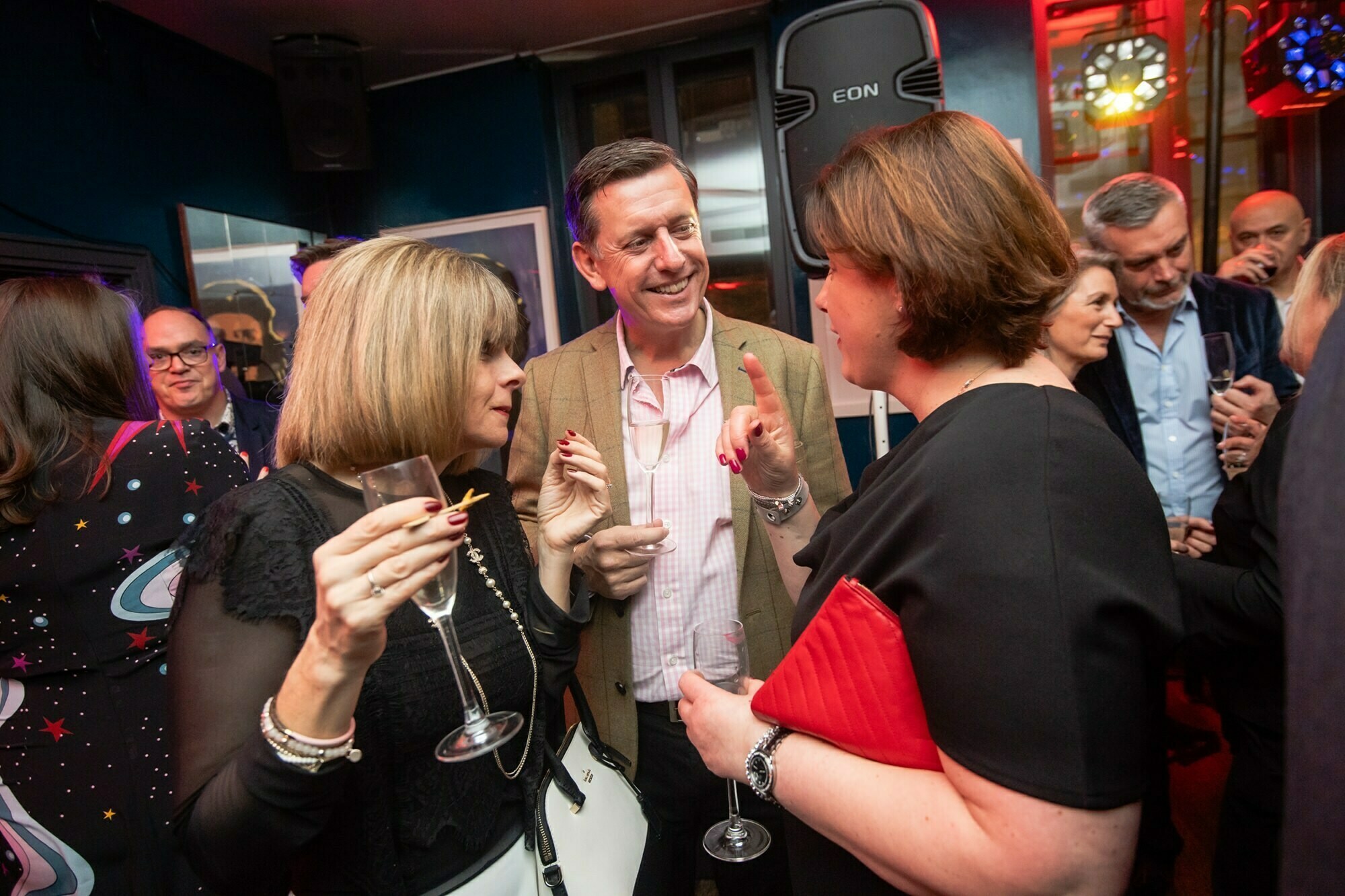 A group of people in formal attire are socializing at an indoor event. They are holding drinks and conversing in a lively atmosphere with dim lighting and colorful decor in the background. James Gifford-Mead Photography - Event Photographer London