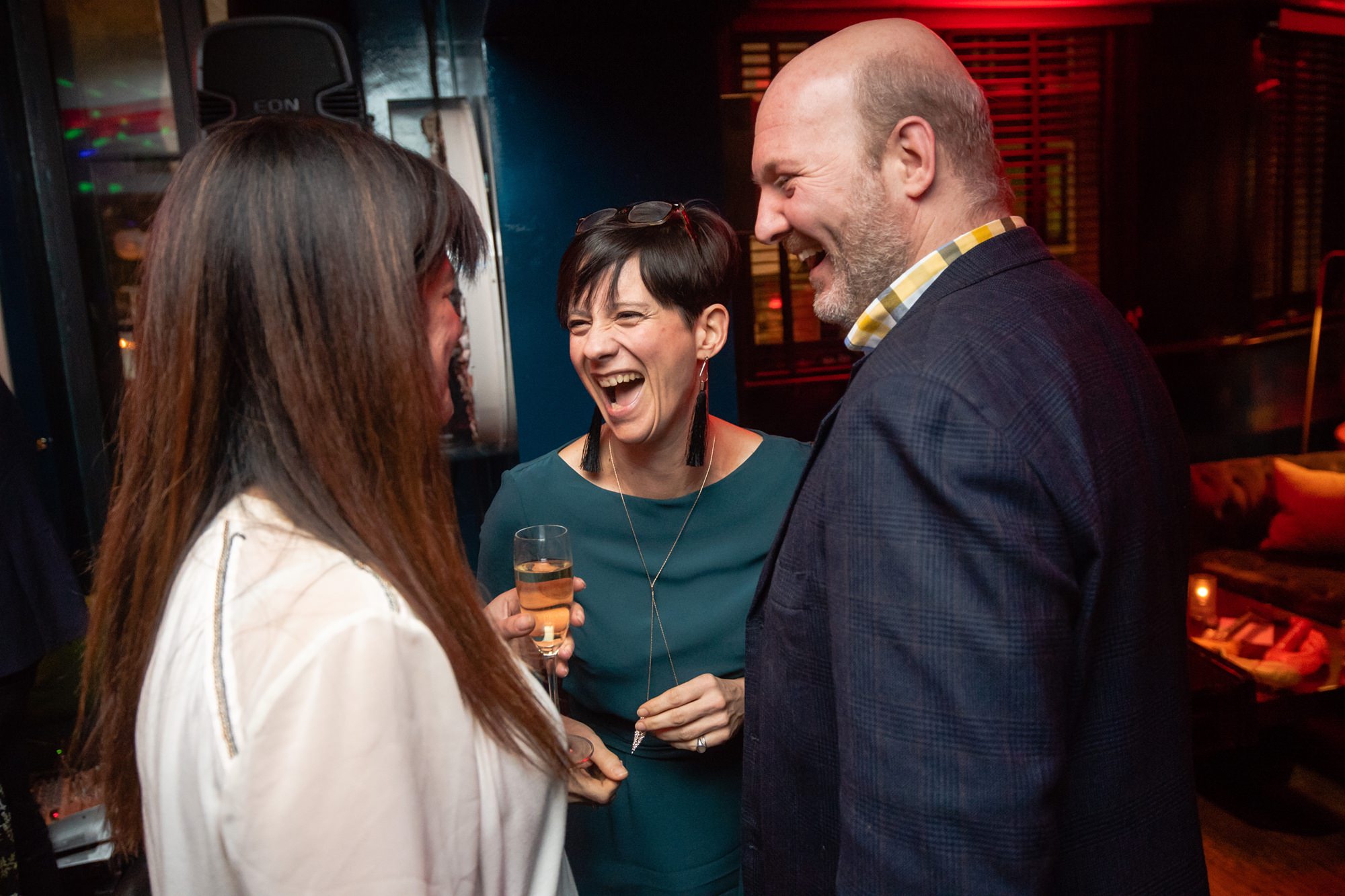 Three people are engaged in conversation at a social gathering. A woman in green is laughing while holding a champagne glass. They appear to be enjoying each other's company in a dimly lit room with warm, ambient lighting. James Gifford-Mead Photography - Event Photographer London