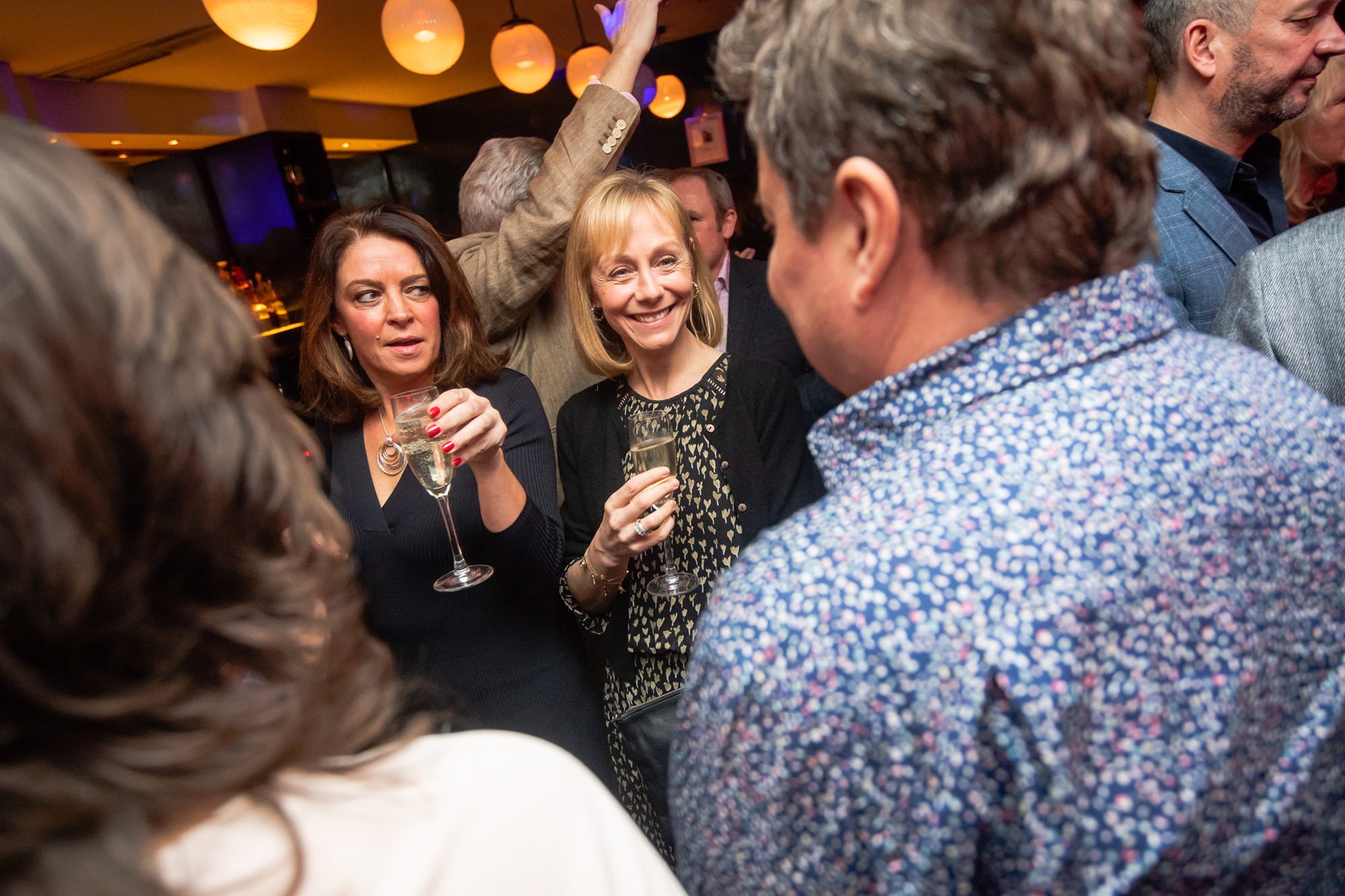 A lively party scene with people socializing. Two women in the center hold glasses and smile, surrounded by others. The background has warm lighting and hanging round lamps, creating a festive atmosphere. James Gifford-Mead Photography - Event Photographer London