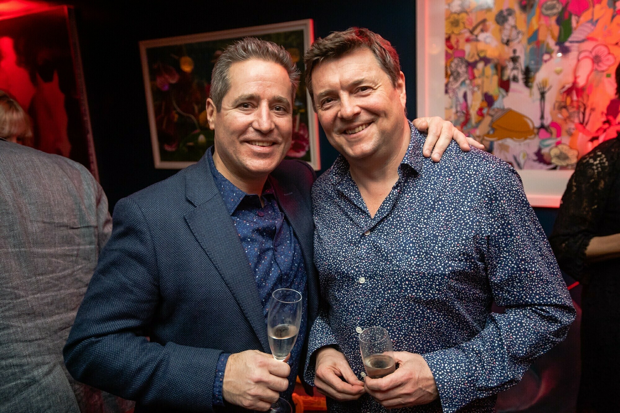Two men are smiling and posing together at an indoor event. Both are holding champagne glasses and wearing patterned shirts. Colorful artwork is visible on the wall behind them, along with a red light casting a warm glow in the room. James Gifford-Mead Photography - Event Photographer London