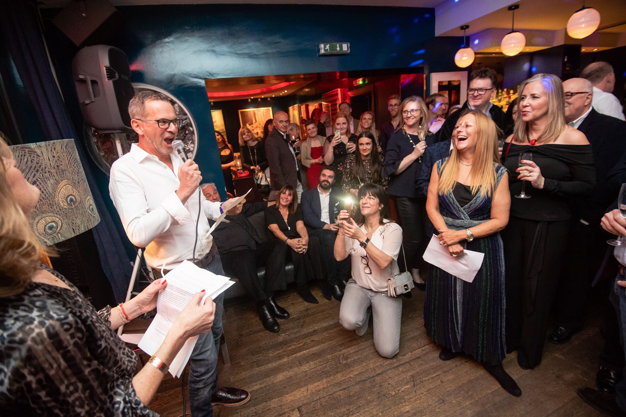 A man with glasses and a microphone speaks to a crowd in a lively room. Several people are gathered, some smiling or holding drinks. A woman kneels, taking photos. The atmosphere is warm and festive, with colorful lighting and wood flooring. James Gifford-Mead Photography - Event Photographer London