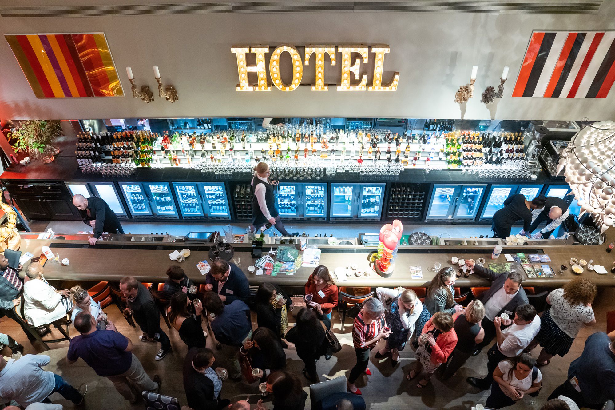 Overhead view of a busy bar in a hotel. Many people are mingling and seated along the bar. A bartender is working, and the word "HOTEL" is illuminated on the wall behind the bar, above a row of bottled drinks and glasses. James Gifford-Mead Photography - Event Photographer London