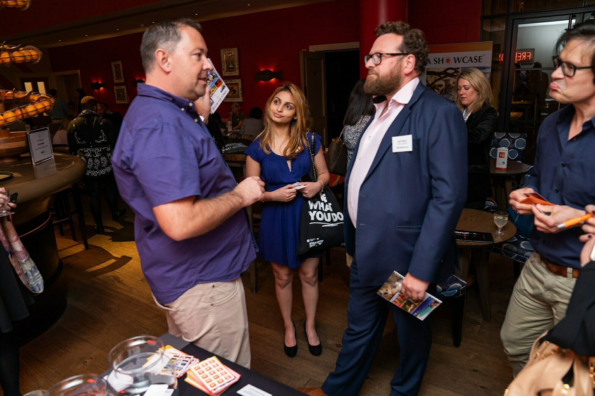 A group of people are gathered in a social setting. One person in a blue shirt speaks while holding a piece of paper. Others listen attentively. A woman in a blue dress stands holding a bag. The room has a warm ambiance with red walls and soft lighting. James Gifford-Mead Photography - Event Photographer London
