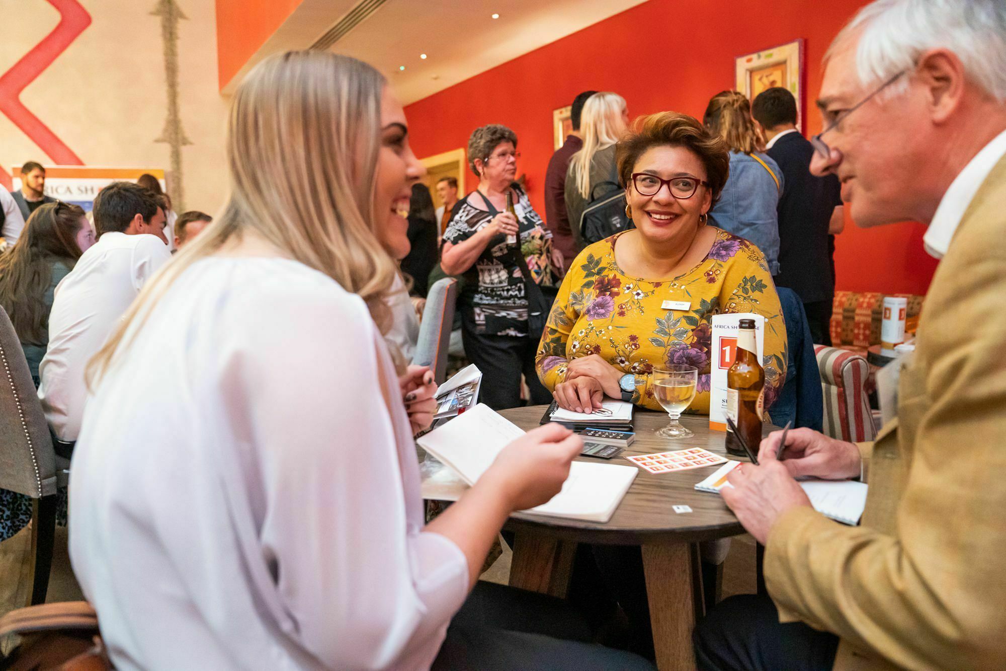 Three people seated at a small round table, engaged in conversation during a lively event. The woman in the center wears glasses and a floral top, smiling while interacting with the others. The background is filled with standing attendees and red decor. James Gifford-Mead Photography - Event Photographer London