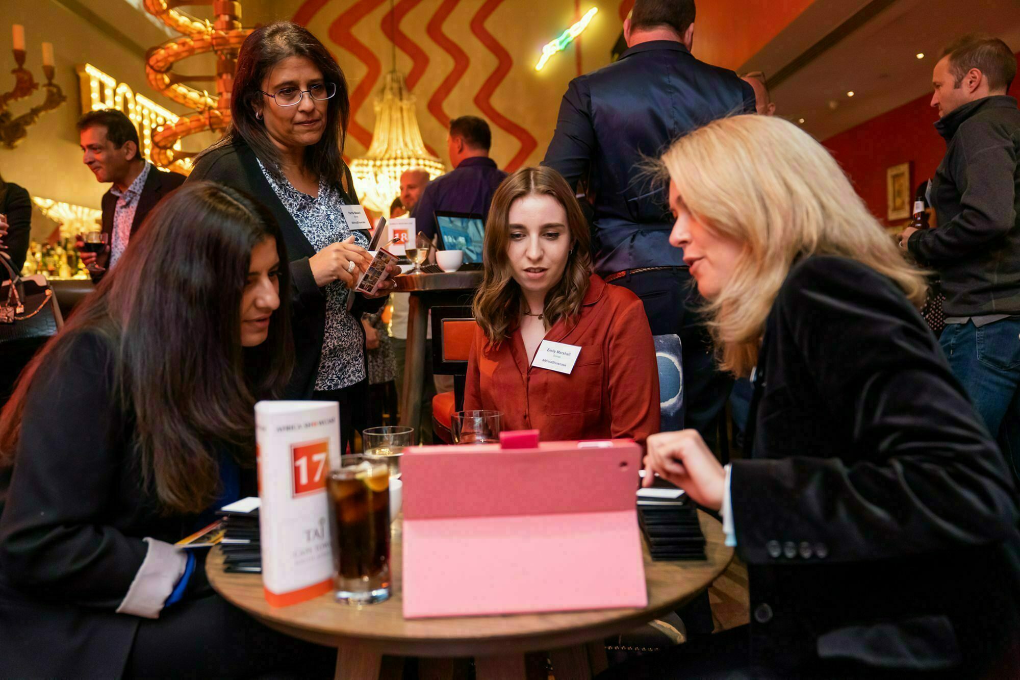 A group of four women sitting around a table in a lively restaurant or event setting, focused on a tablet device. Other people are mingling in the background. The table has drinks and a card with the number 17 on it. James Gifford-Mead Photography - Event Photographer London