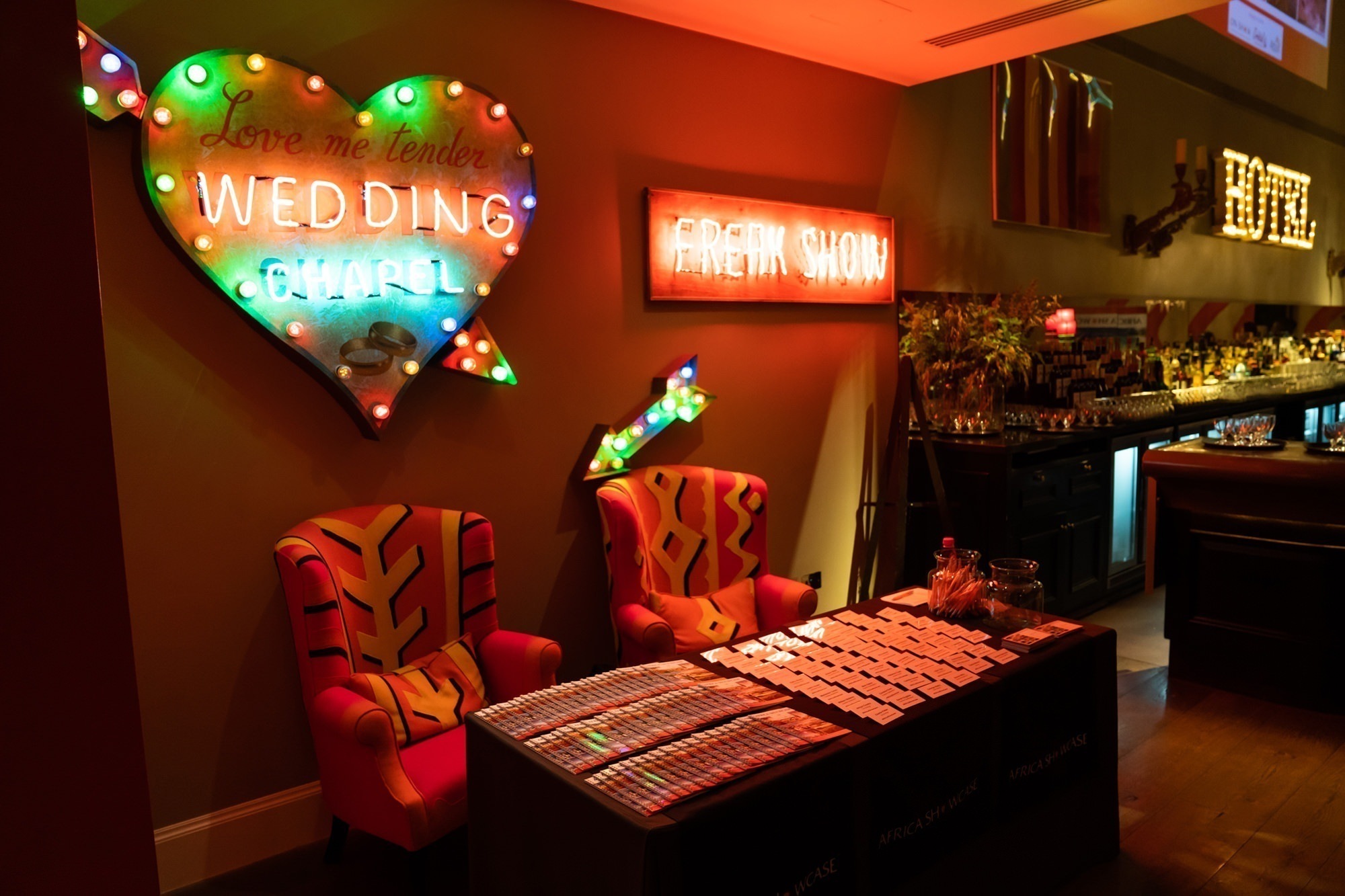 A dimly lit room features a vibrant, heart-shaped neon sign reading "Love me tender, Wedding Chapel." Beside it, "Freak Show" glows in neon. Two red patterned chairs flank a table covered with papers. A bar area is visible in the background. James Gifford-Mead Photography - Event Photographer London