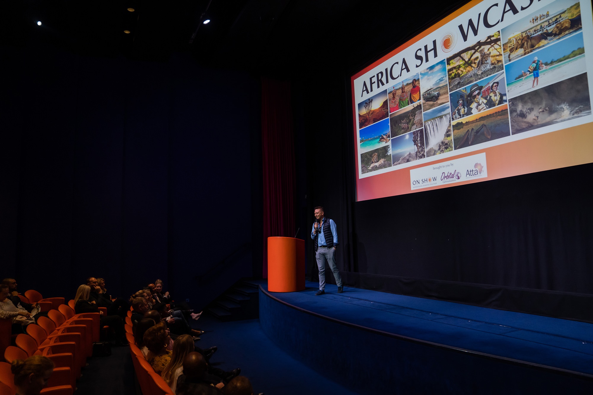 Man standing on stage behind a podium, speaking to an audience in a theater. A screen displays images of landscapes and wildlife with the title "Africa Showcase." Rows of orange seats are partially visible. James Gifford-Mead Photography - Event Photographer London