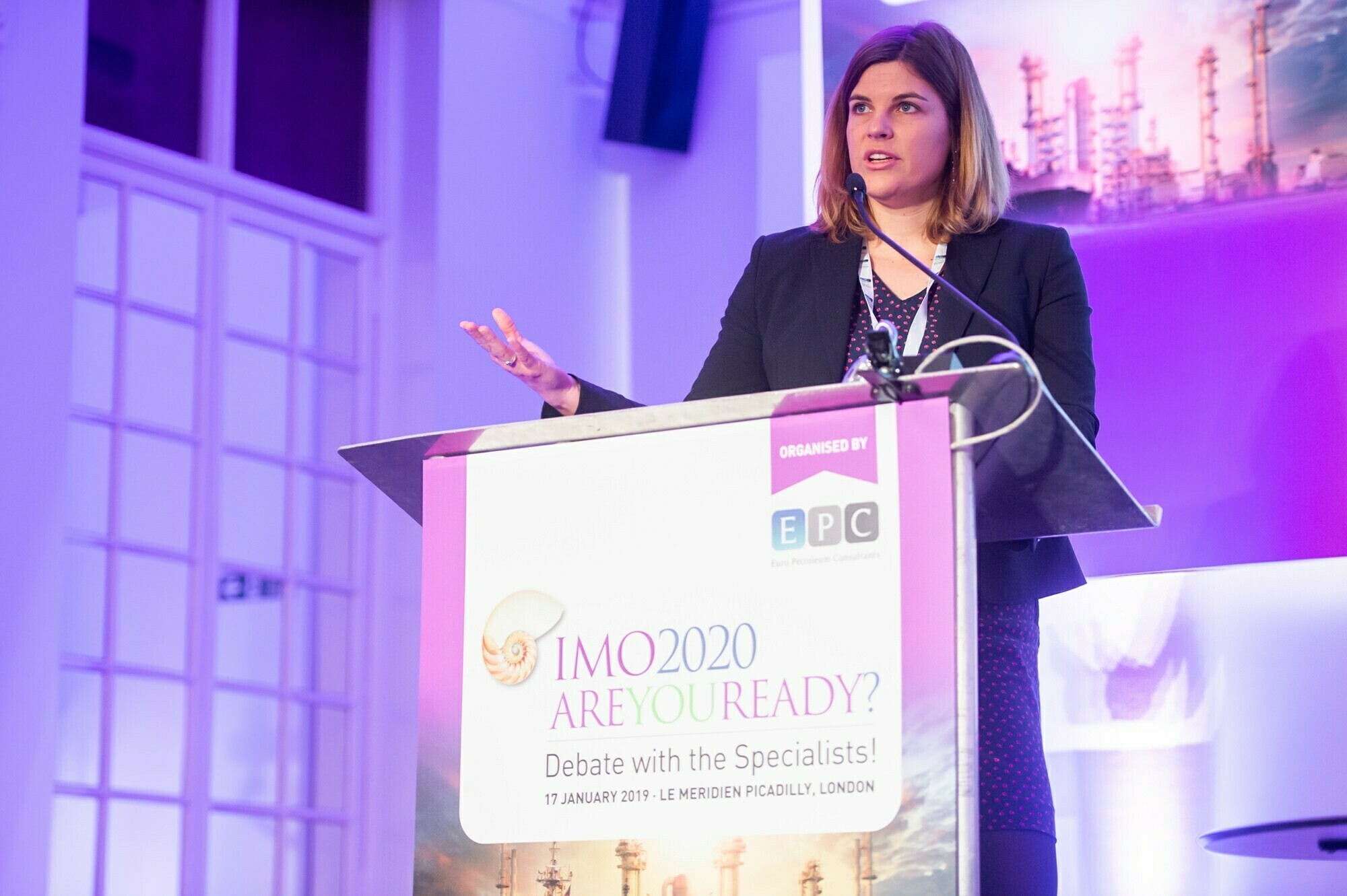 A woman stands at a podium speaking, with a microphone attached to her. The banner on the podium reads, "IMO 2020 Are You Ready? Debate with the Specialists," dated 17 January 2019, at Le Meridien Piccadilly, London.