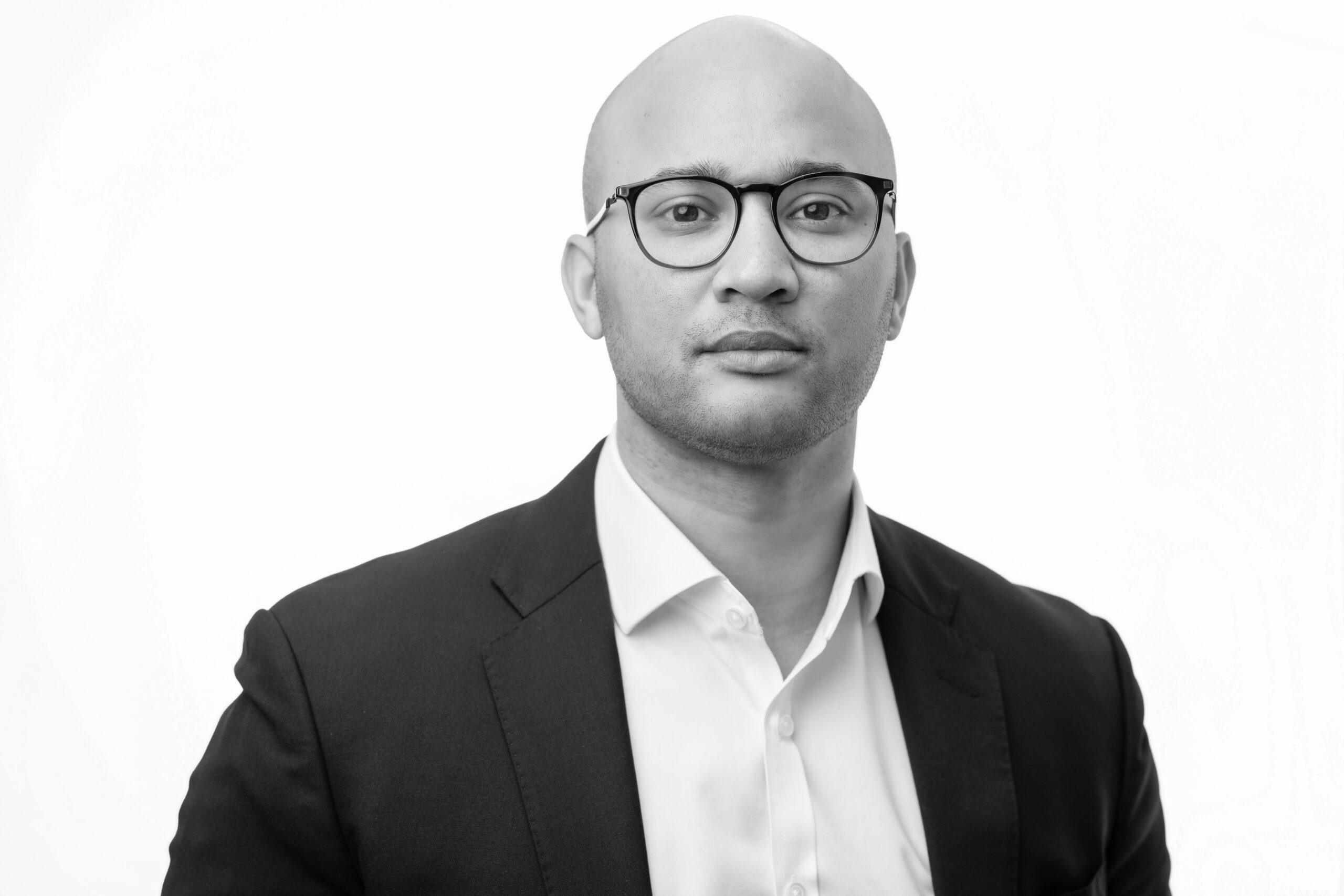 A person in a black suit and white shirt is looking straight ahead. They are wearing glasses and have a neutral expression. The background is white. The image is in black and white. James Gifford-Mead Photography - Event Photographer London
