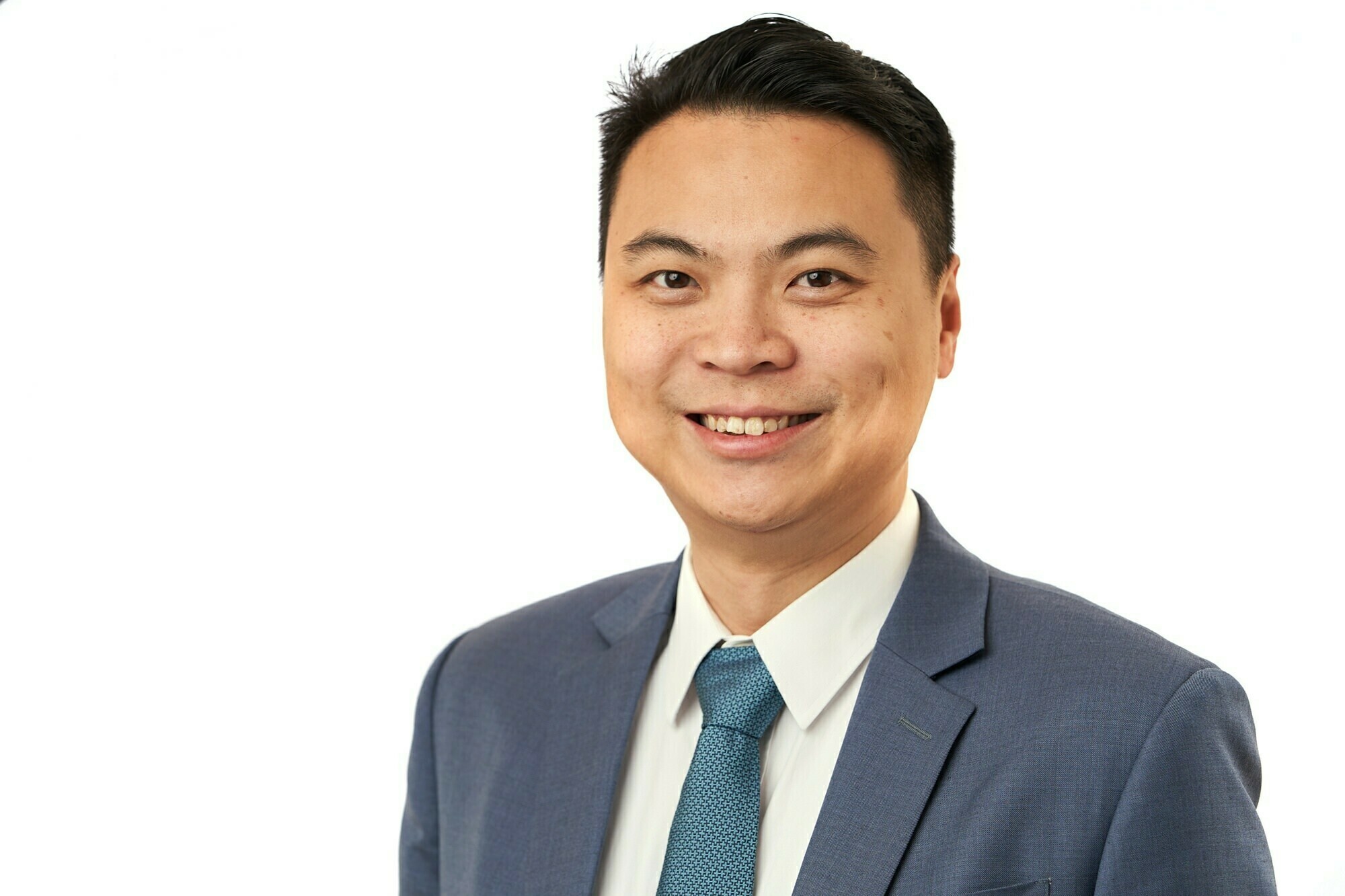 A man in a blue suit and tie smiles at the camera. The background is plain white, creating a professional and clean look. James Gifford-Mead Photography - Event Photographer London