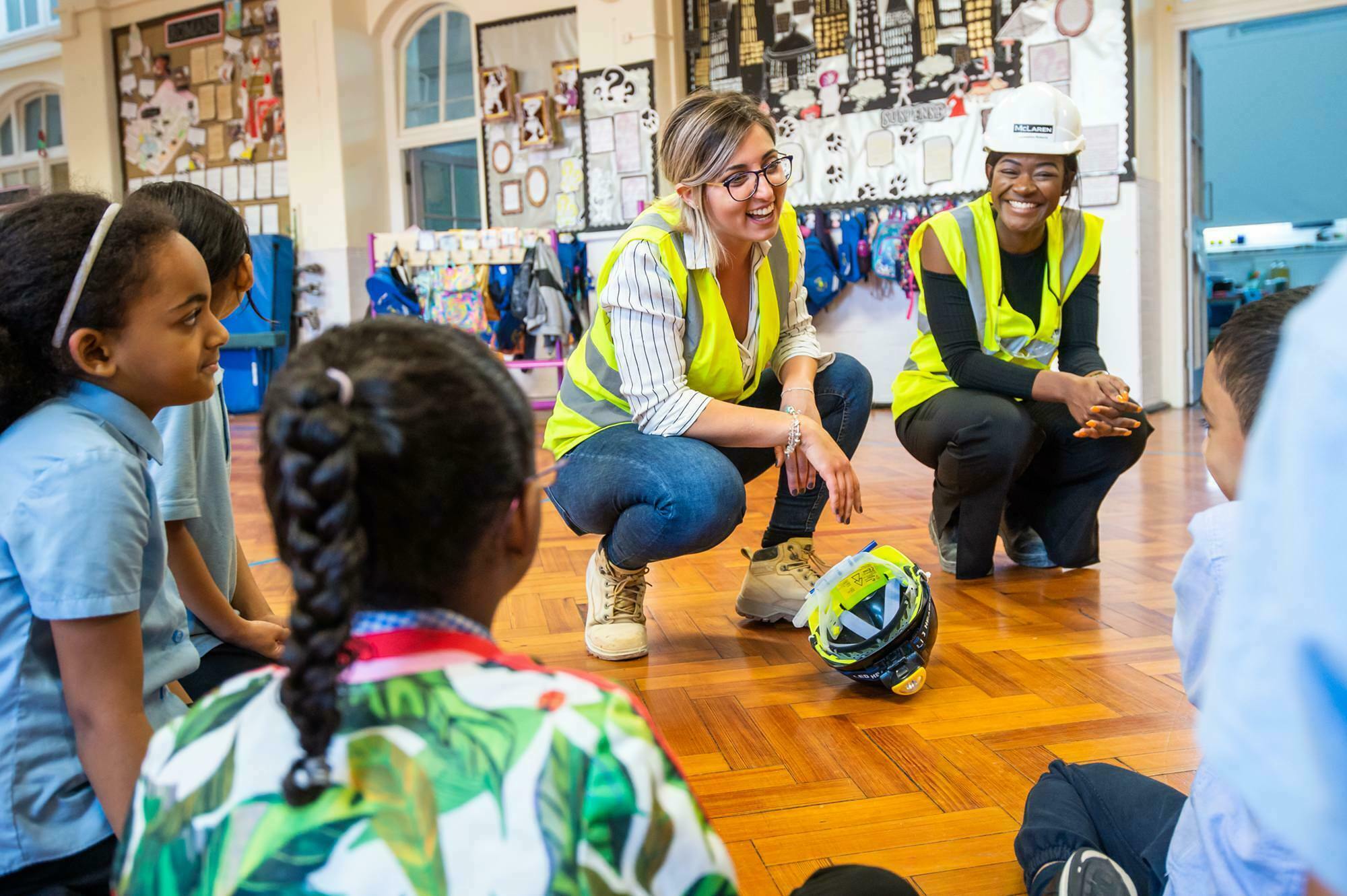 Two women wearing safety vests and a hard hat are talking to seated children in a classroom. They are crouching on a wooden floor, smiling, with one helmet placed on the floor. The background includes colorful decorations and clothing hooks. James Gifford-Mead Photography - Event Photographer London