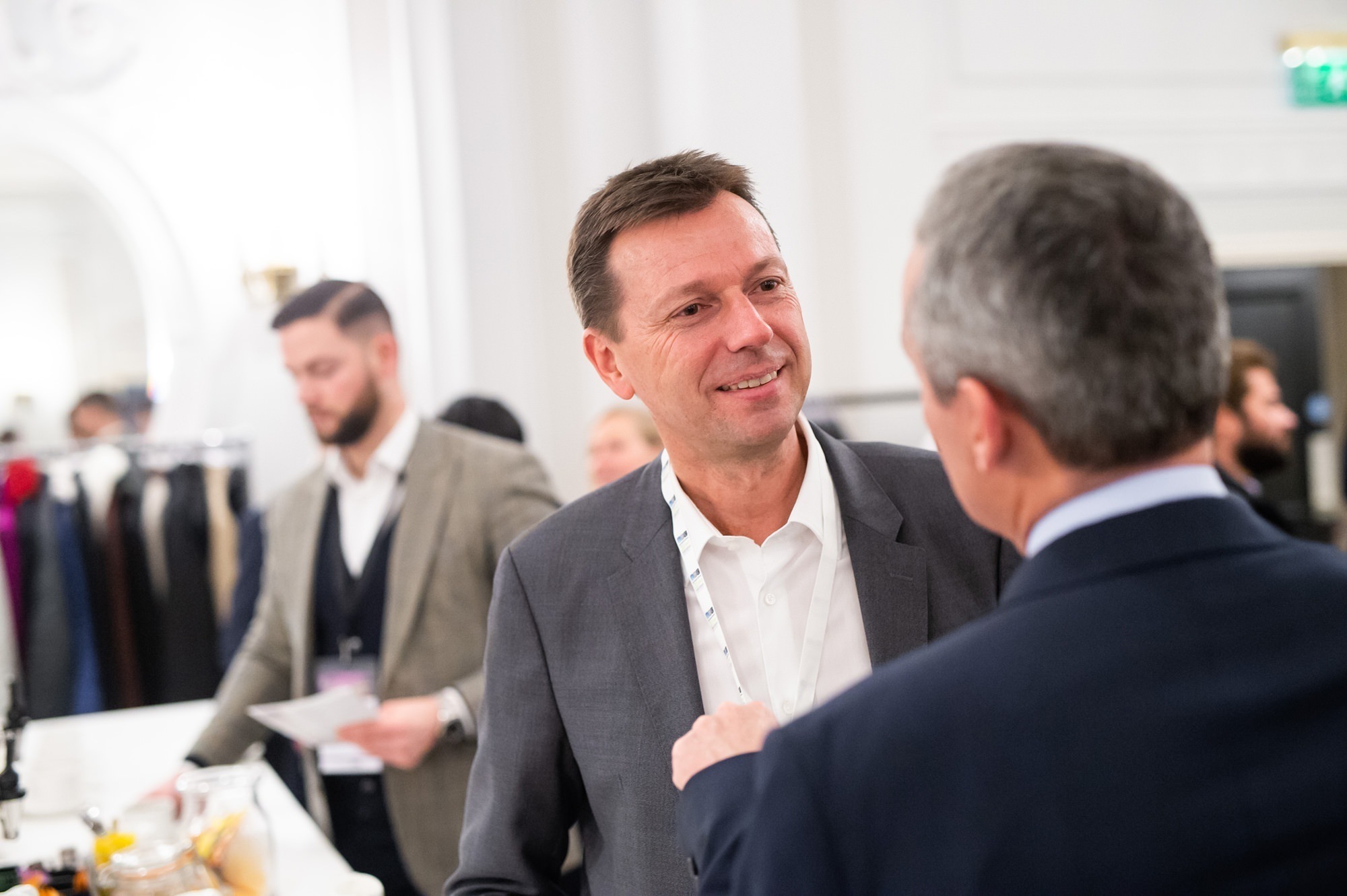 Two men in suits are engaged in conversation in a well-lit room. One man has a lanyard around his neck. A blurred figure in the background stands near a rack of clothes. The setting appears formal, suggesting a business or networking event. James Gifford-Mead Photography - Event Photographer London