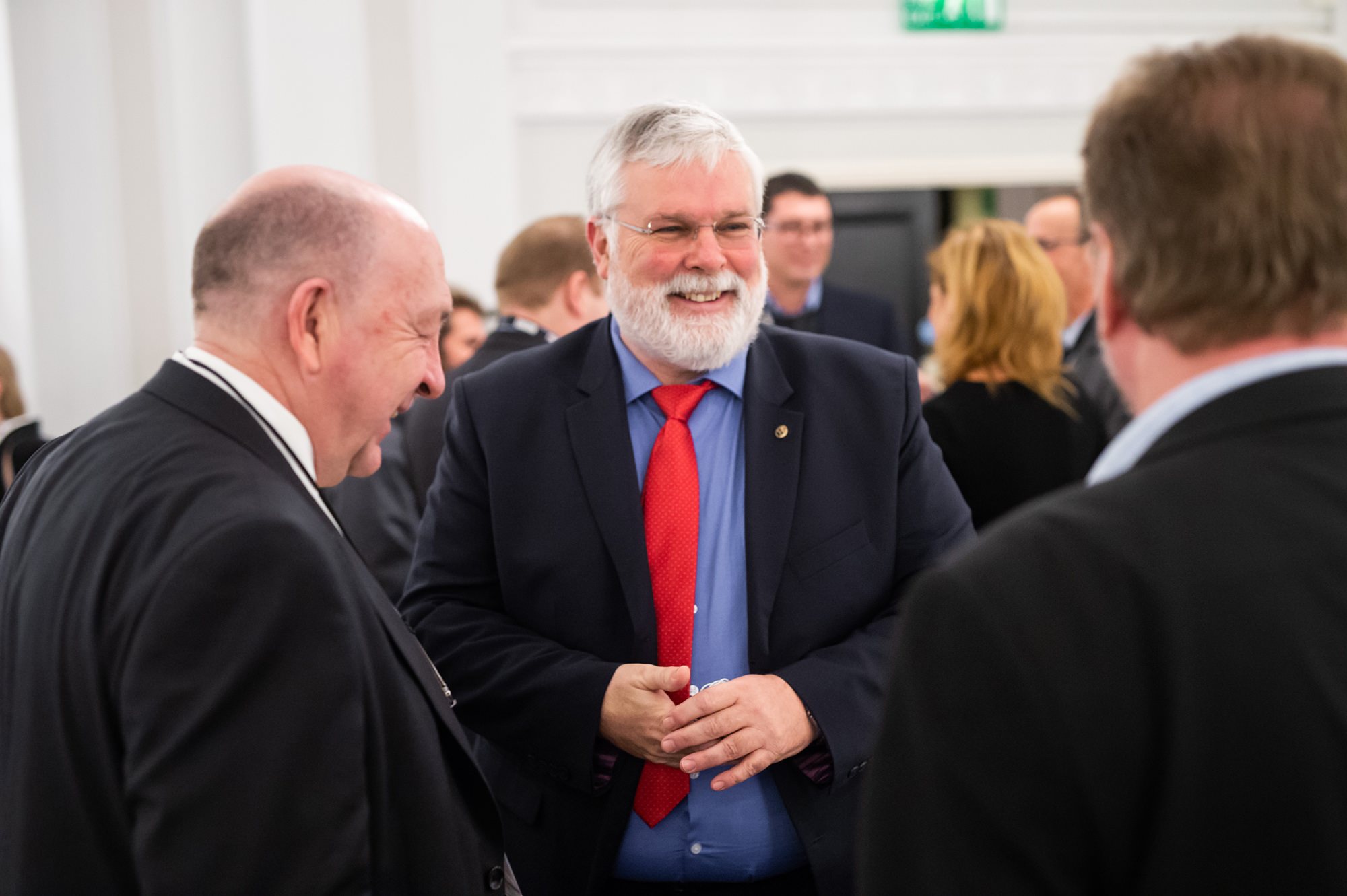 A group of people in formal attire are engaged in conversation indoors. A man with a white beard and red tie is smiling at two other men, one bald and the other with short brown hair. The setting appears to be a conference or meeting. James Gifford-Mead Photography - Event Photographer London
