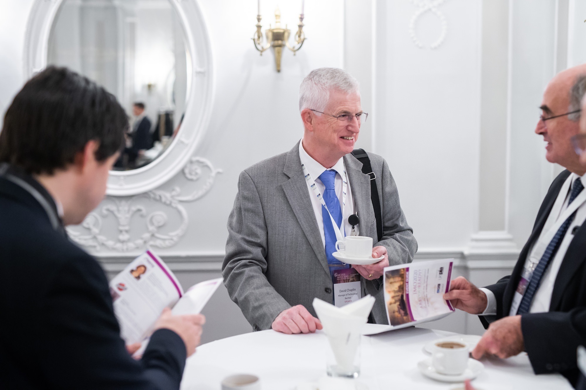 Three men in business attire are standing at a round table, engaged in conversation. One holds a cup, while two others look at brochures. The setting appears to be a formal event or conference, with elegant white walls and a mirror in the background. James Gifford-Mead Photography - Event Photographer London