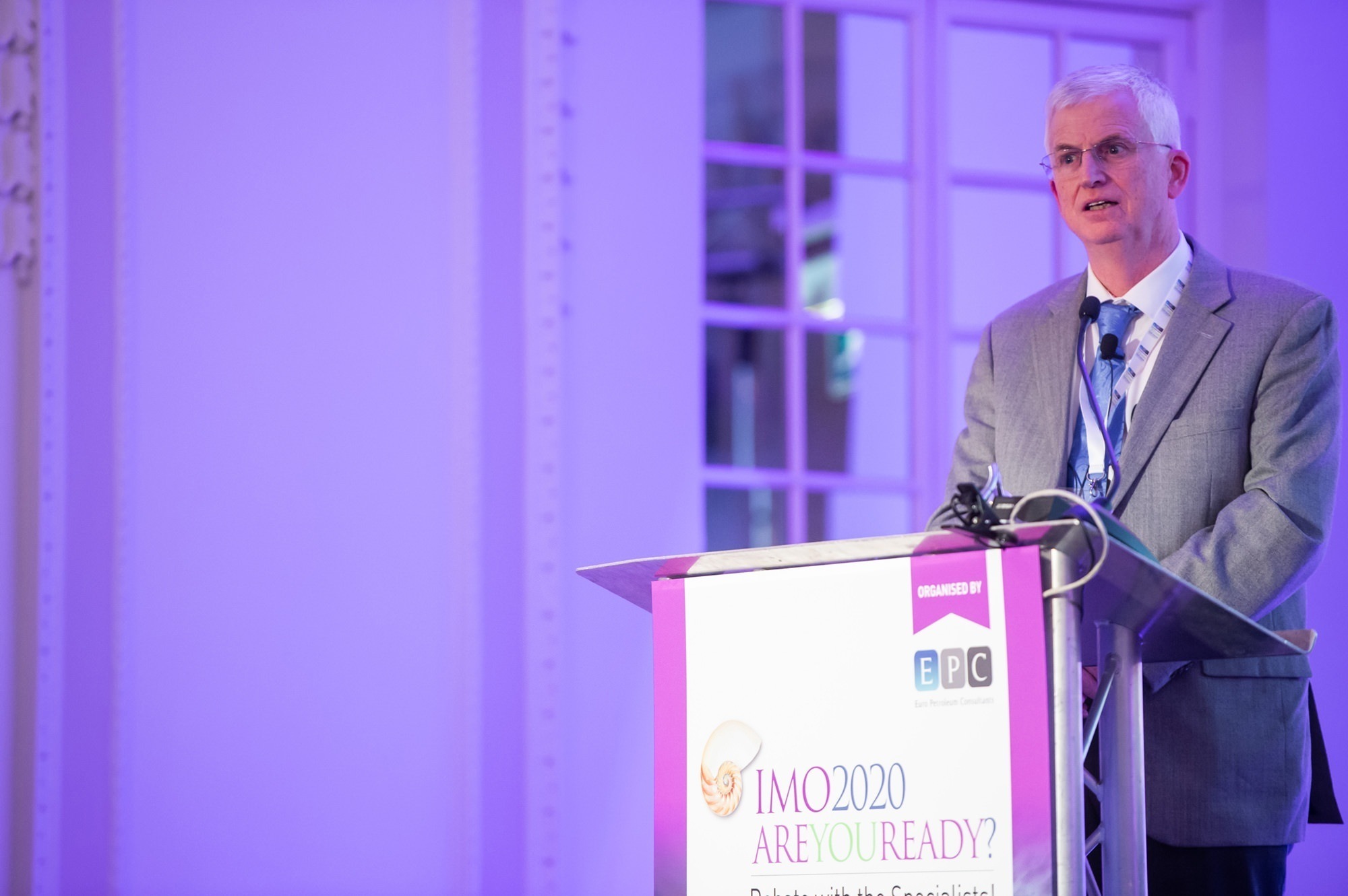A man in a gray suit and glasses speaks at a podium with a microphone. The podium displays a sign reading "IMO 2020 Are You Ready?" The background is purple-lit with windows. James Gifford-Mead Photography - Event Photographer London