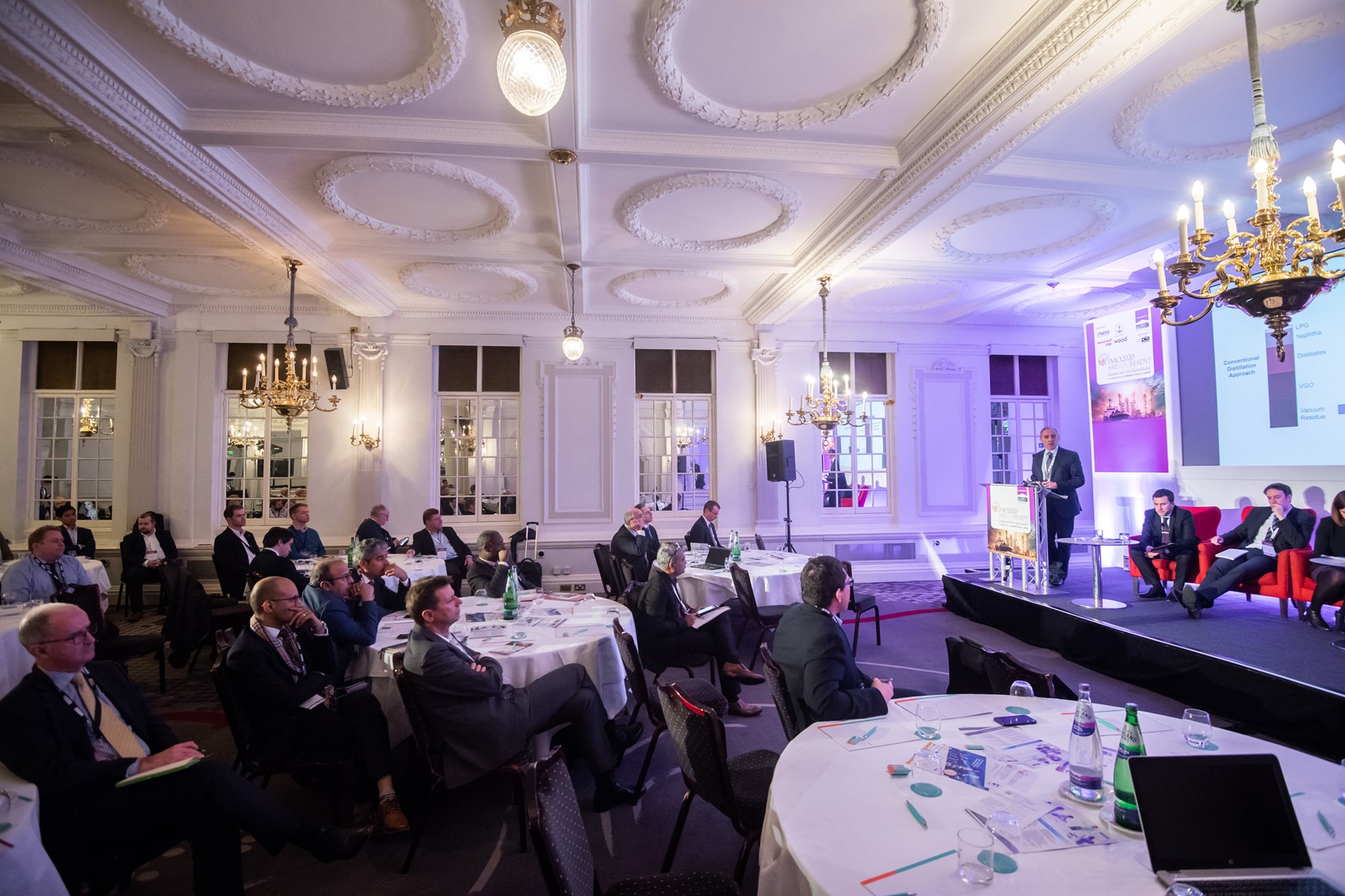 A conference room filled with people seated at round tables, listening to a speaker at a podium. The room has elegant chandeliers and ornate ceiling designs. Presentation slides are visible on a screen, and attendees are taking notes. James Gifford-Mead Photography - Event Photographer London