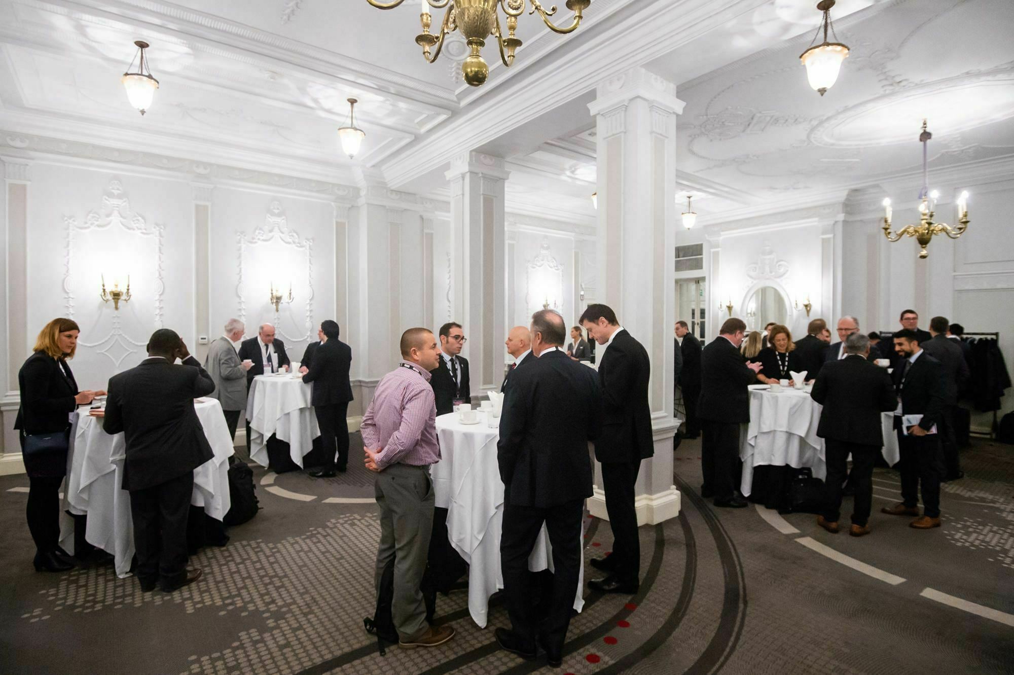 People in formal attire are mingling at a networking event in an elegant room with chandeliers and white columns. They stand and talk around several high tables covered with white tablecloths. James Gifford-Mead Photography - Event Photographer London