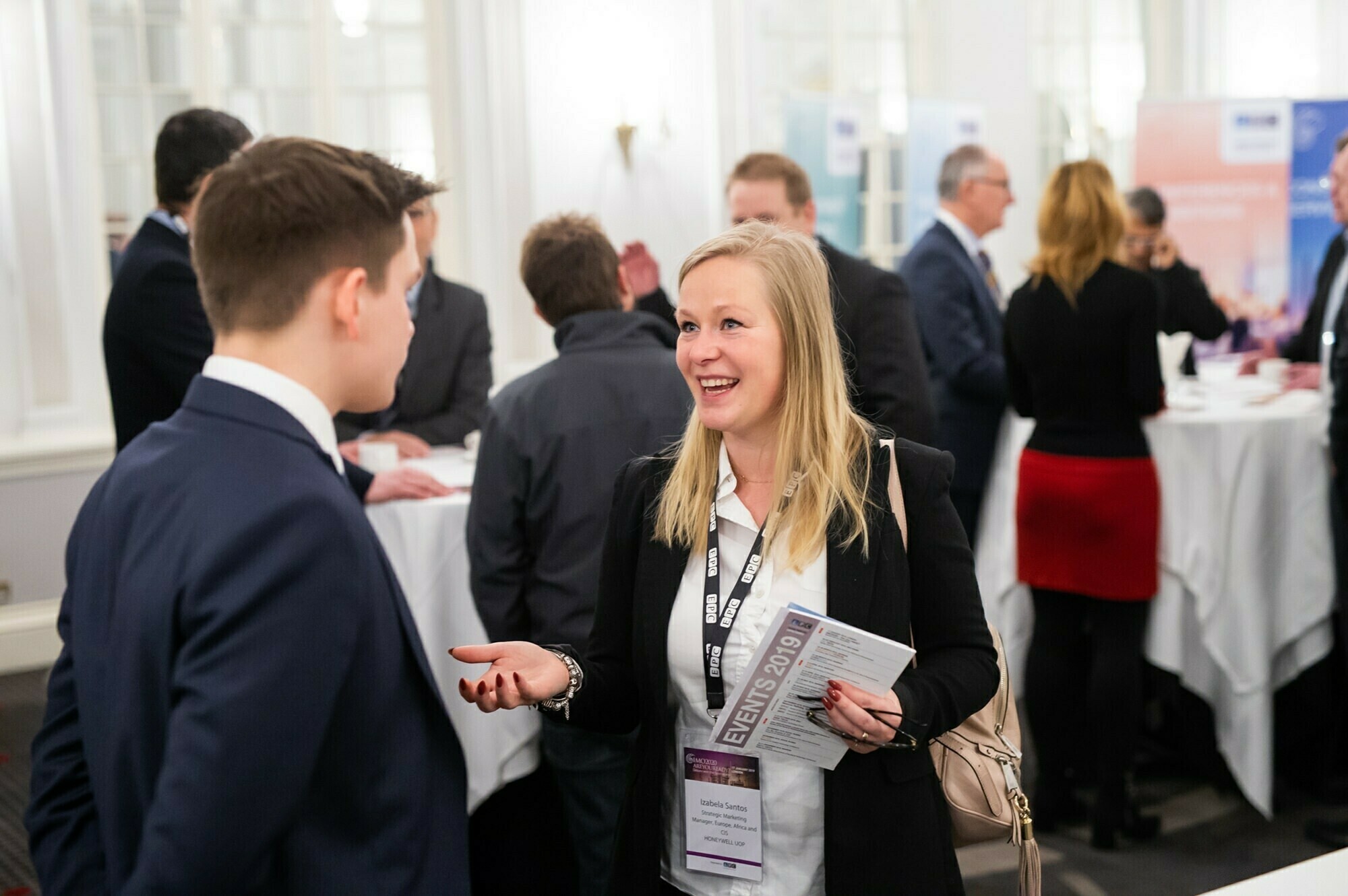 A woman in conversation with a man at a professional networking event. She holds a pamphlet while other attendees engage in discussions in the background. Tables with white tablecloths are visible. James Gifford-Mead Photography - Event Photographer London