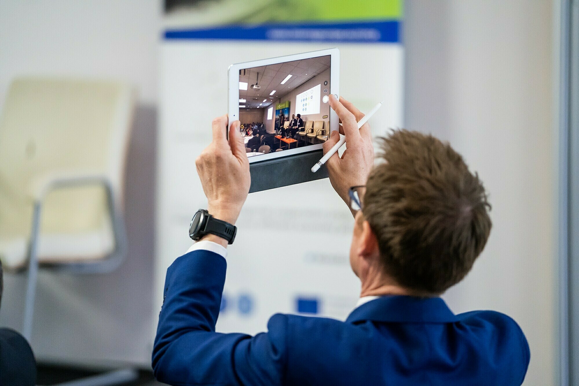 A man in a blue suit holds a white tablet and a stylus, taking a photo of a conference or presentation. A blurred screen and chairs are visible on the tablet's display. An empty chair is in the background. James Gifford-Mead Photography - Event Photographer London
