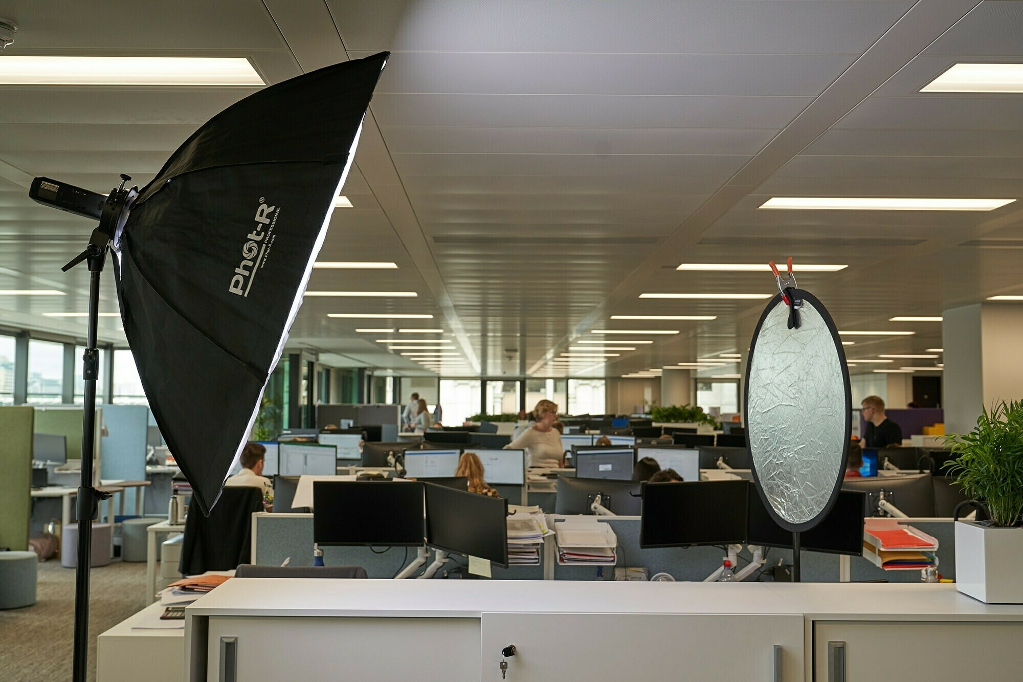 A modern office with rows of desks and computers. A large photography light and a reflector are set up in the foreground, indicating a photoshoot or video recording session. The ceiling lights illuminate the spacious area. James Gifford-Mead Photography - Event Photographer London