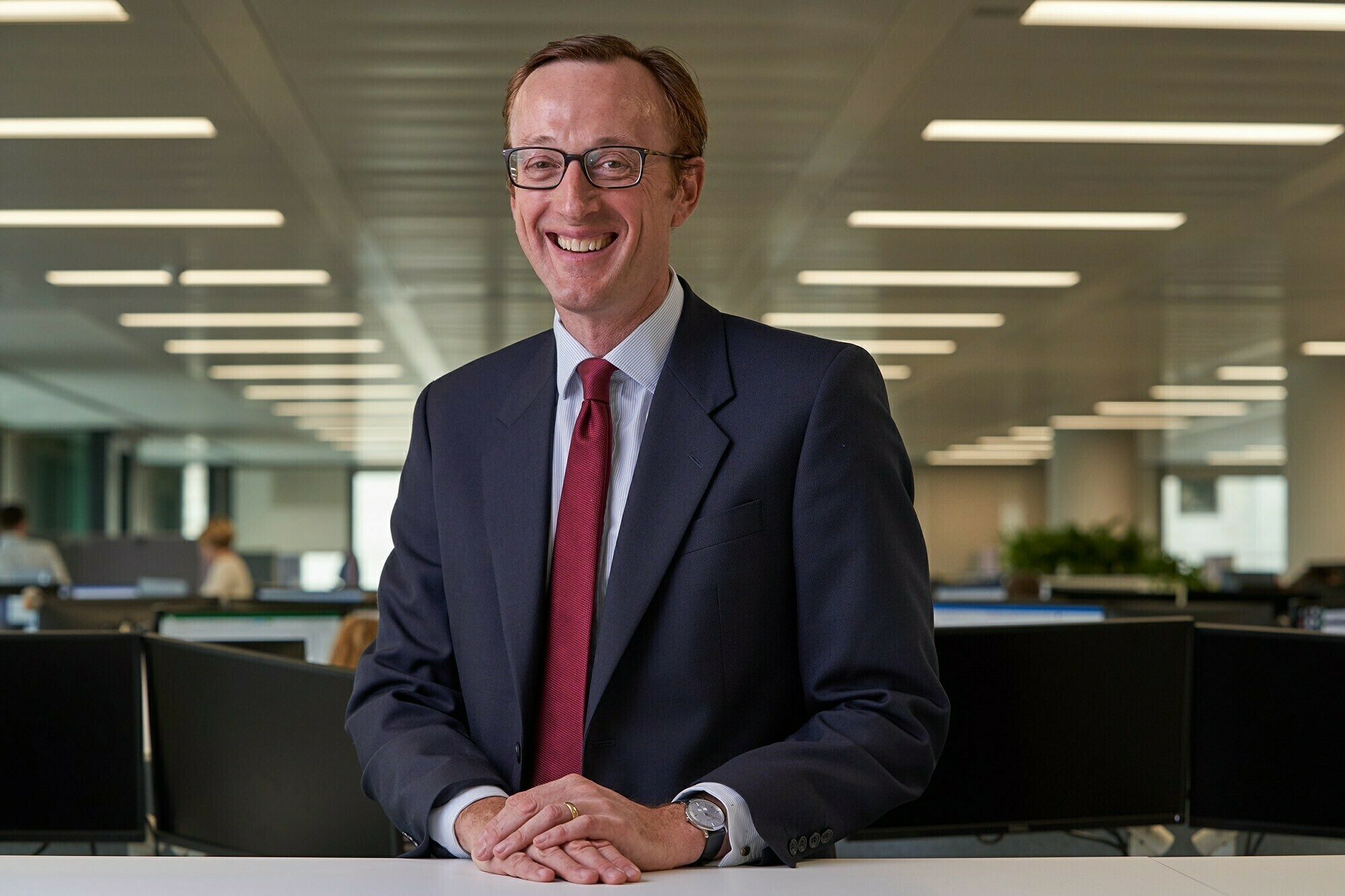 A smiling man in a suit and red tie stands in a modern office with fluorescent lighting. He wears glasses and a watch, and the background features open workspaces with several people in the distance. James Gifford-Mead Photography - Event Photographer London