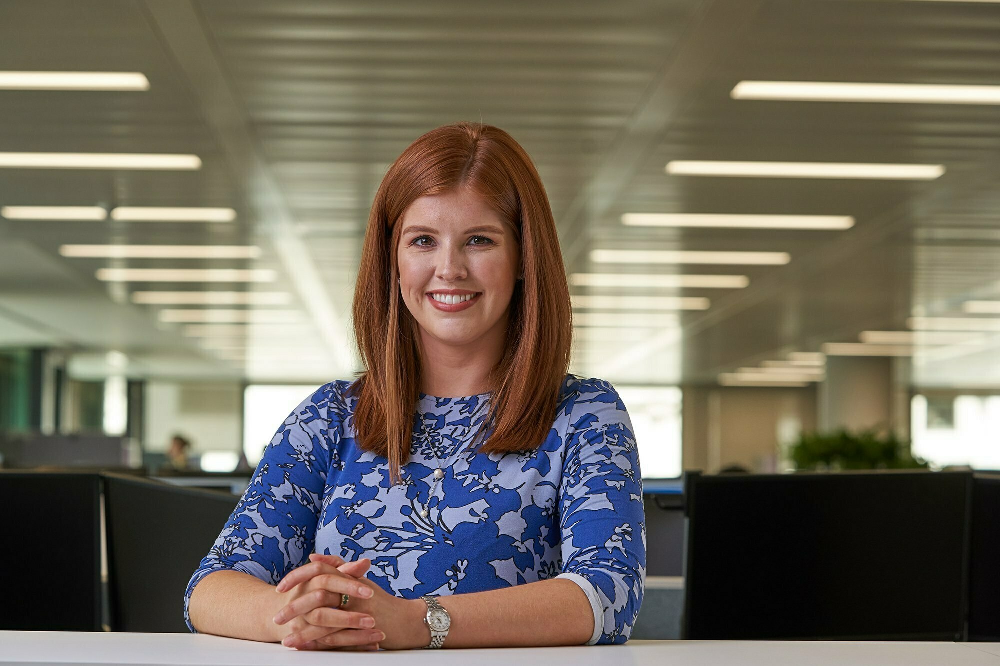 A woman with long red hair sits at a table in a bright office, smiling at the camera. She wears a blue floral-patterned top and a watch. The background features rows of lights and office spaces. James Gifford-Mead Photography - Event Photographer London