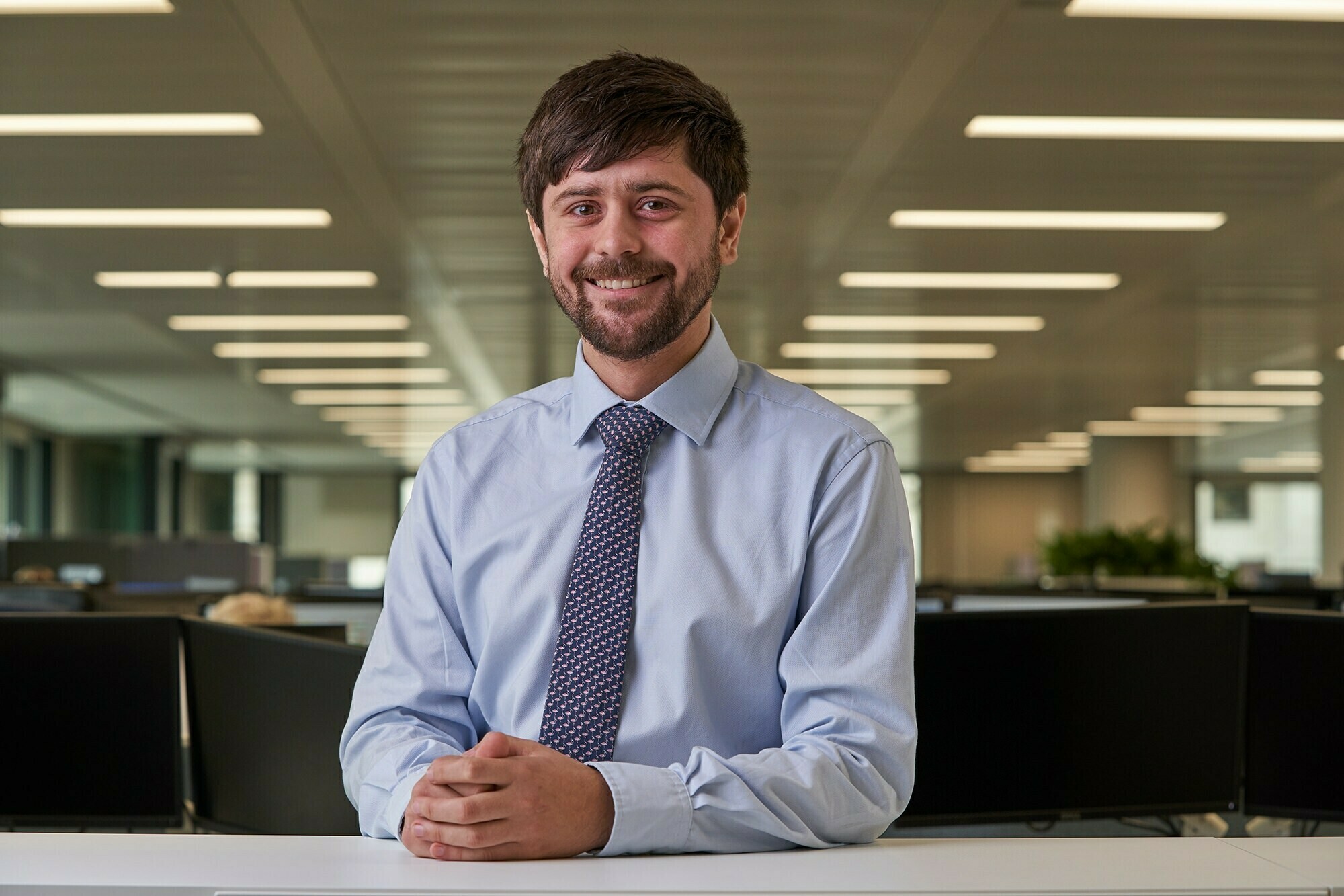 Smiling man with a beard and short hair, wearing a light blue shirt and tie, sits at a desk in an office with a row of lights on the ceiling and several computer monitors in the background. James Gifford-Mead Photography - Event Photographer London