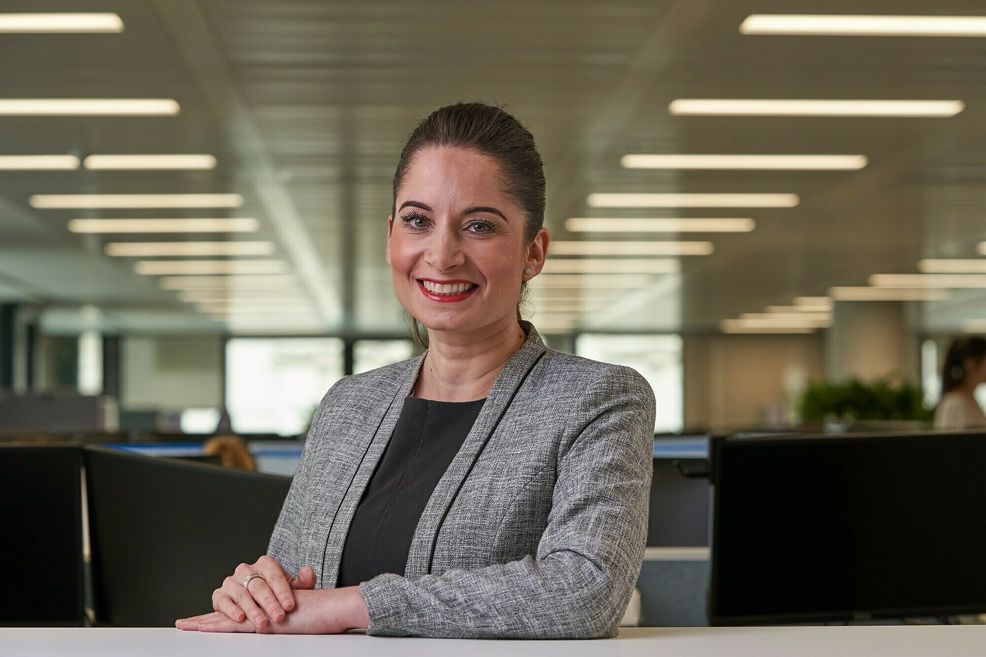 A woman in a gray blazer and black top smiles confidently while standing in a modern office setting. She is posed with her arms crossed, and the background shows a spacious, well-lit area with multiple workstations. James Gifford-Mead Photography - Event Photographer London
