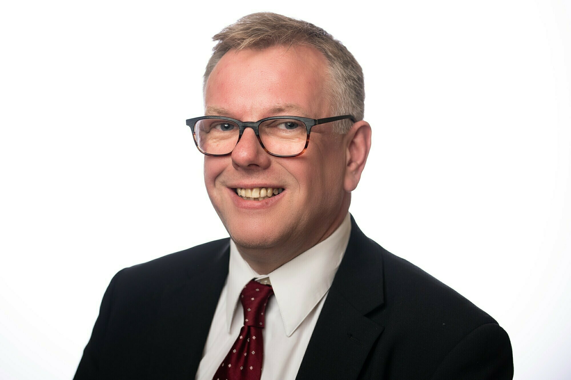 A person with short, light hair and glasses is smiling, wearing a black suit, white shirt, and red polka dot tie. The background is plain white. James Gifford-Mead Photography - Event Photographer London