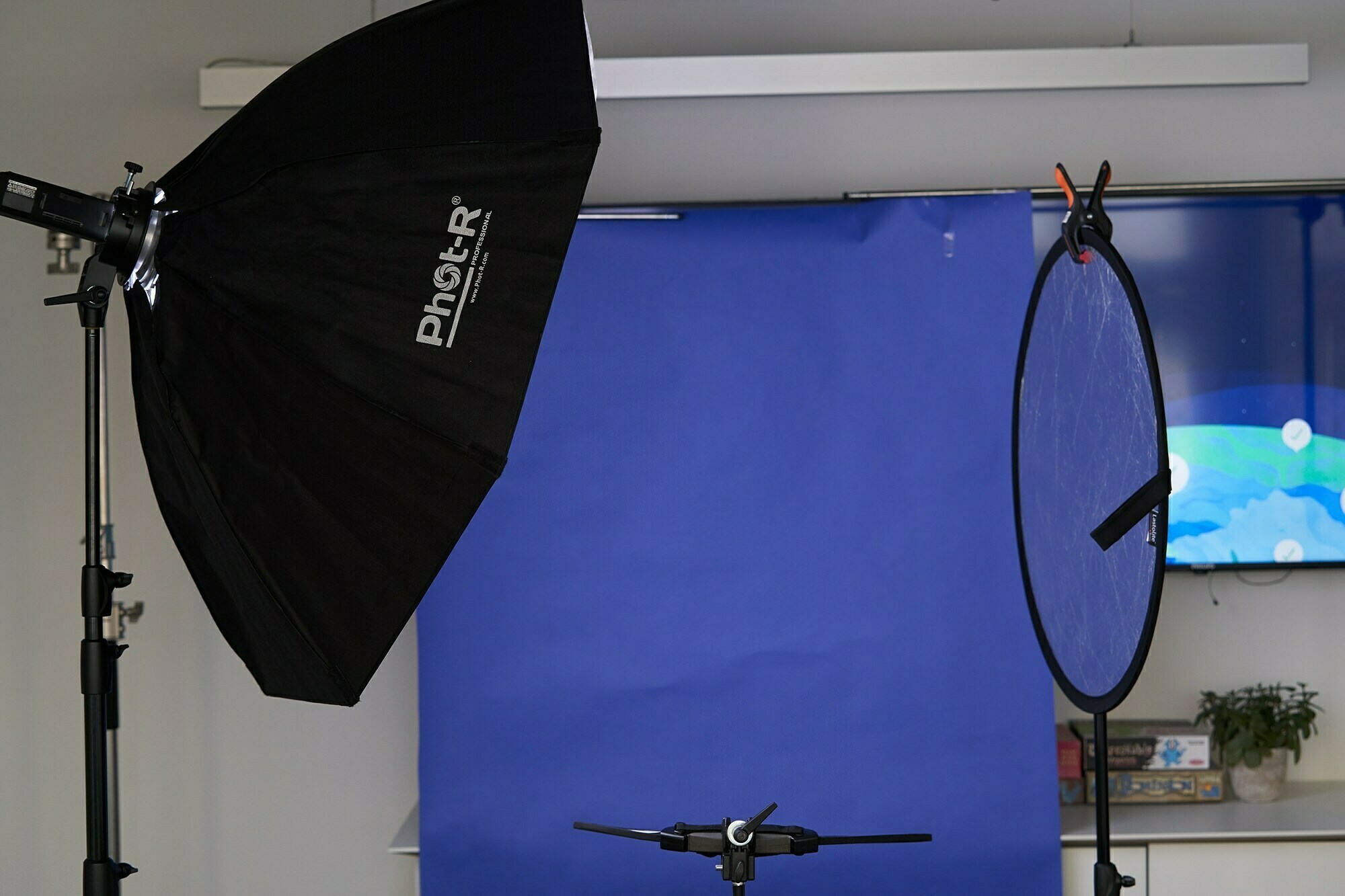 Photography studio setup with a large softbox light on the left, a blue backdrop in the center, and a circular reflector on a stand to the right. Various equipment and a screen with abstract visuals are visible in the background. James Gifford-Mead Photography - Event Photographer London