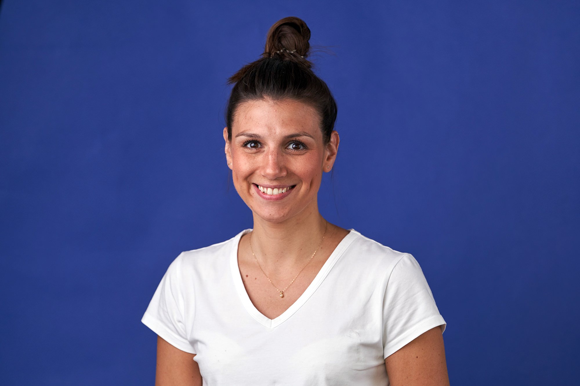 A person with long brown hair in a bun is smiling while wearing a white shirt with a gold necklace. The background is solid dark blue. James Gifford-Mead Photography - Event Photographer London