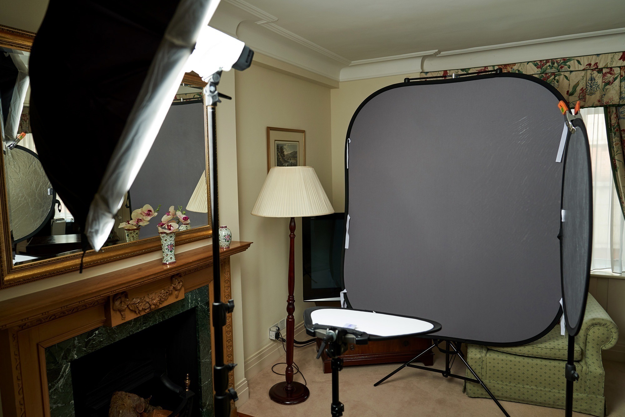 A room is set up for a photo shoot, featuring softbox lights, a large collapsible backdrop, and a small reflector. There is a fireplace with a mirror above it and a lamp near the window with floral curtains. James Gifford-Mead Photography - Event Photographer London