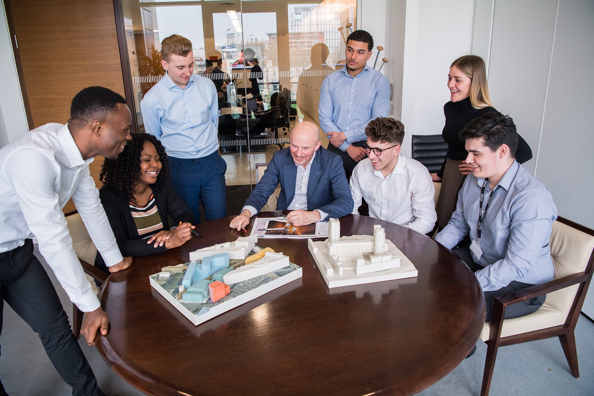 A diverse group of eight people gather around a table in an office, examining architectural models. They are smiling, engaged in discussion. A cityscape is visible through the window in the background. James Gifford-Mead Photography - Event Photographer London