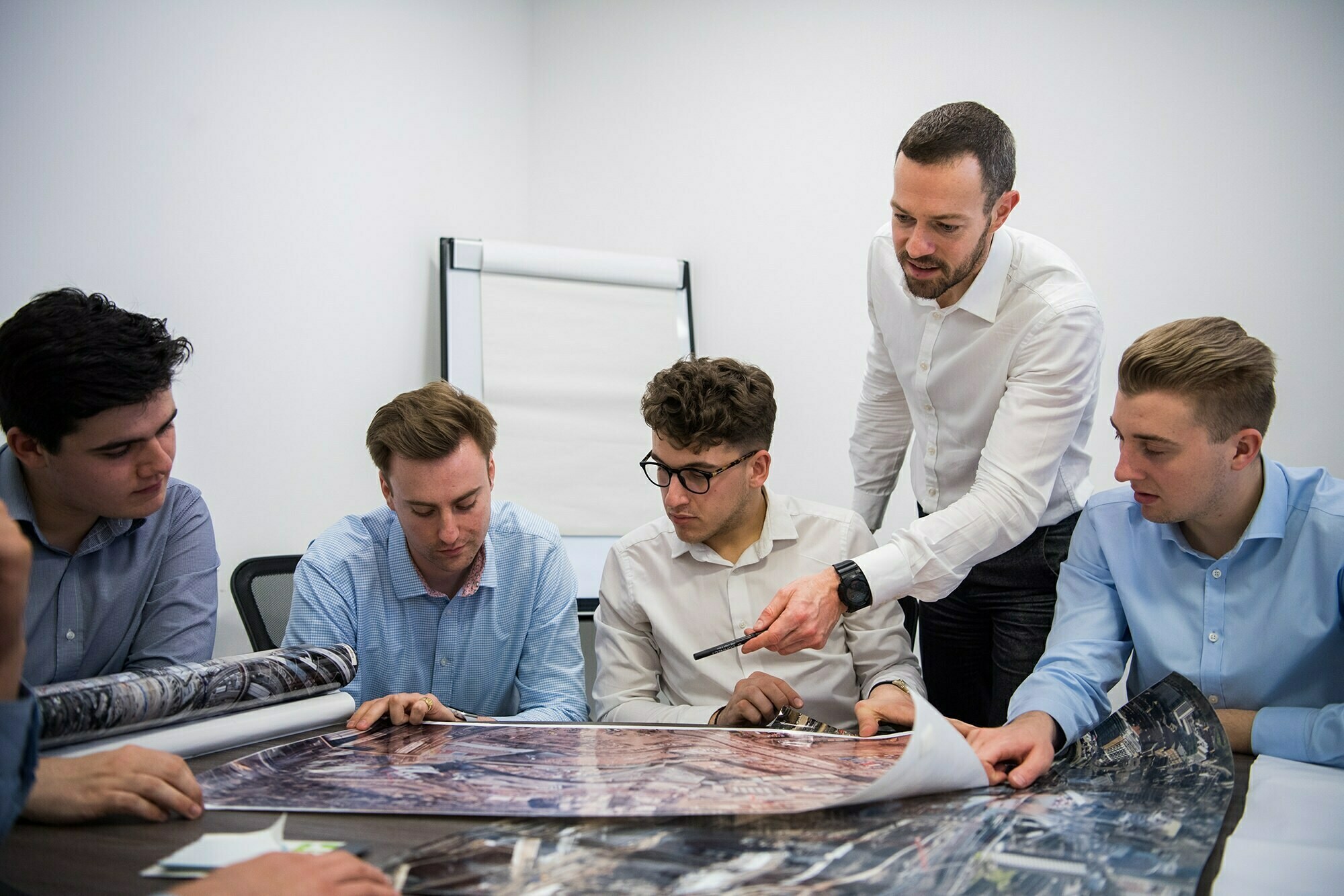 A group of five men gathered around a table, discussing plans. They are examining a large, printed map or blueprint. One man, standing, is pointing at the map with a pen. An easel with a blank pad is in the background. James Gifford-Mead Photography - Event Photographer London