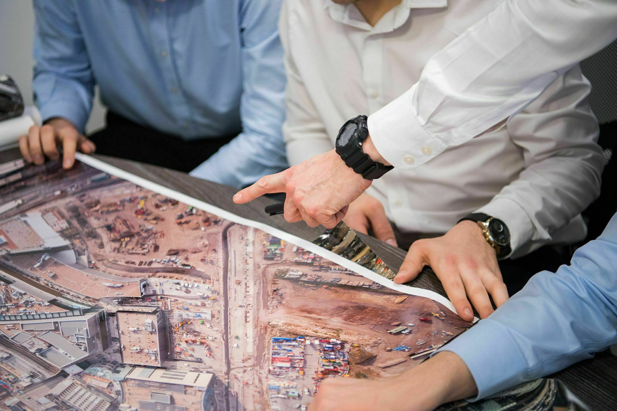 Three people in business attire lean over a large aerial photo of a construction site. One person is pointing at the image, discussing details with colleagues who are closely examining the plans. James Gifford-Mead Photography - Event Photographer London