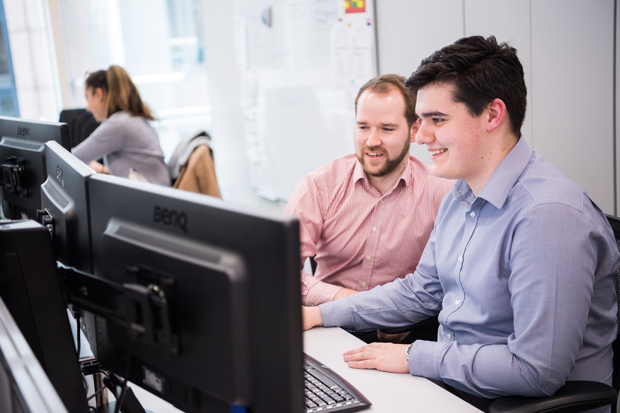 Two men in a modern office work on a computer together, smiling and focused on the screen. They are dressed in business casual attire. In the background, another person is working at a desk. James Gifford-Mead Photography - Event Photographer London