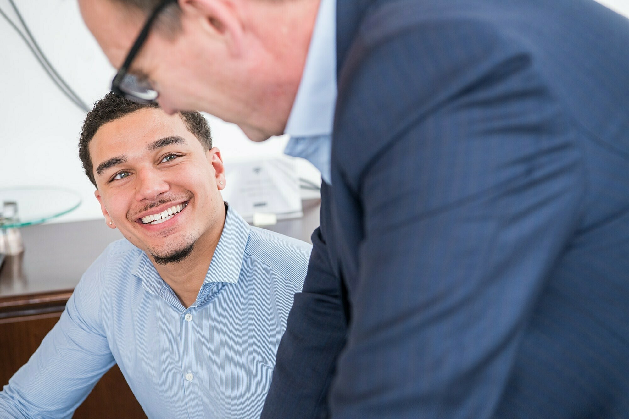 A young man in a light blue shirt smiles while looking up at another person in a dark suit. They appear to be in an office setting, with a desk and papers in the background. James Gifford-Mead Photography - Event Photographer London
