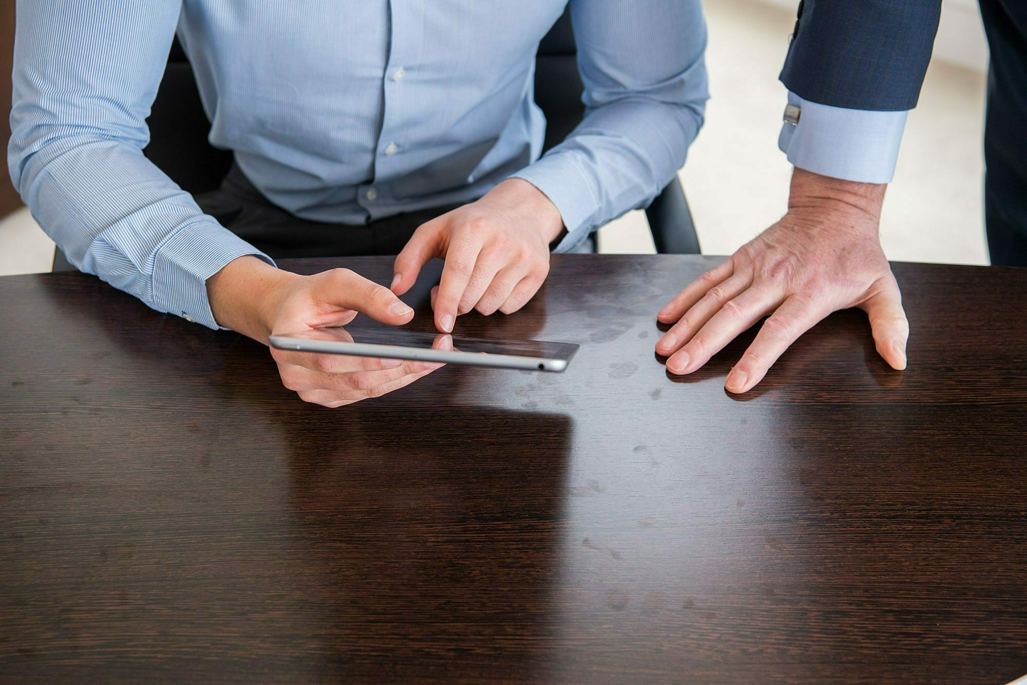 Two people are at a wooden table. One person, seated, is using a tablet, their finger pointing at the screen. The other person, standing, has their hand placed on the table. Both are wearing business attire. James Gifford-Mead Photography - Event Photographer London