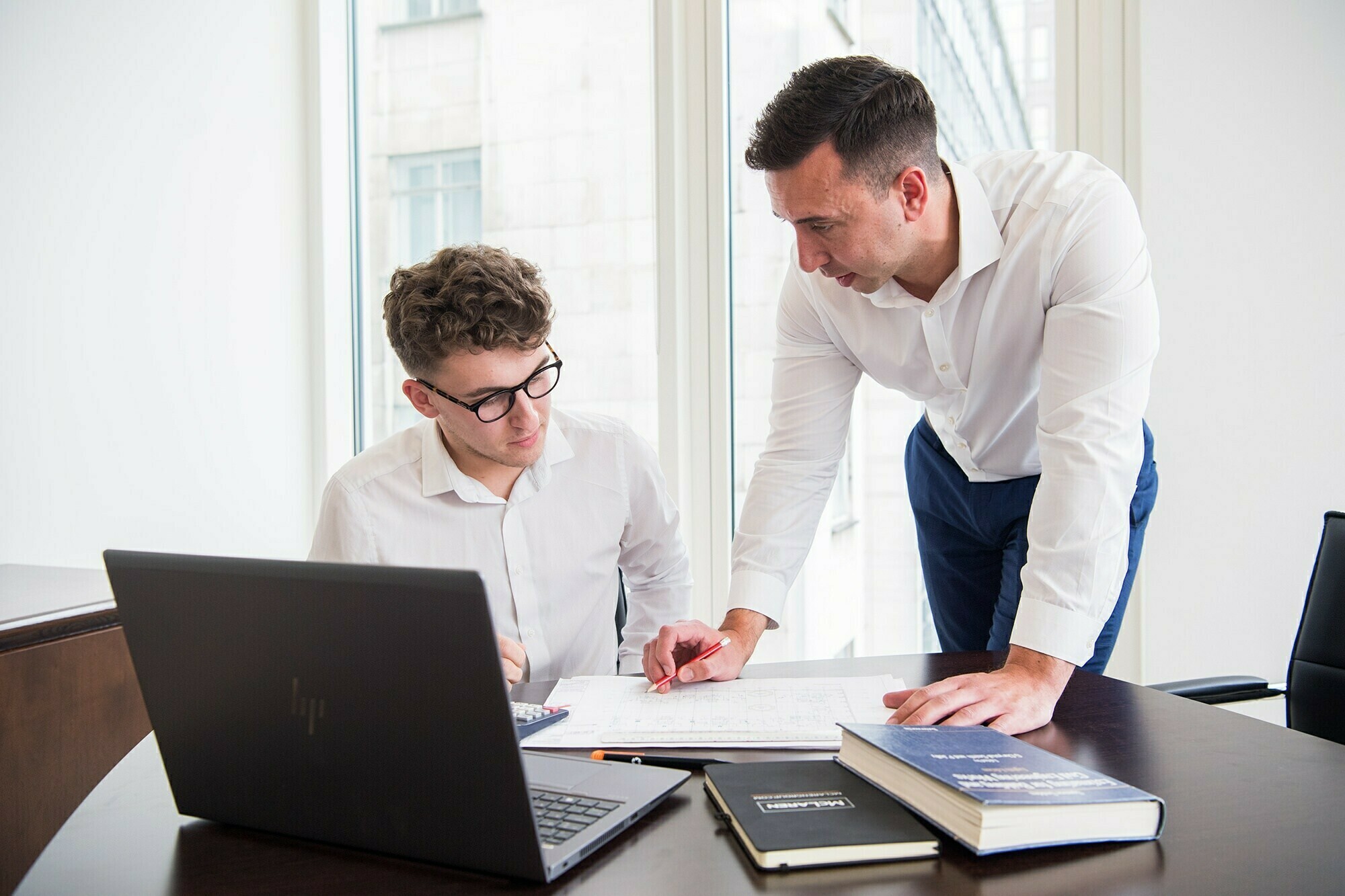 Two men in an office, one seated at a desk with a laptop and the other standing, discussing over a notebook. Both are wearing white shirts. A book and a notepad are also on the table near them. Large windows provide natural light. James Gifford-Mead Photography - Event Photographer London