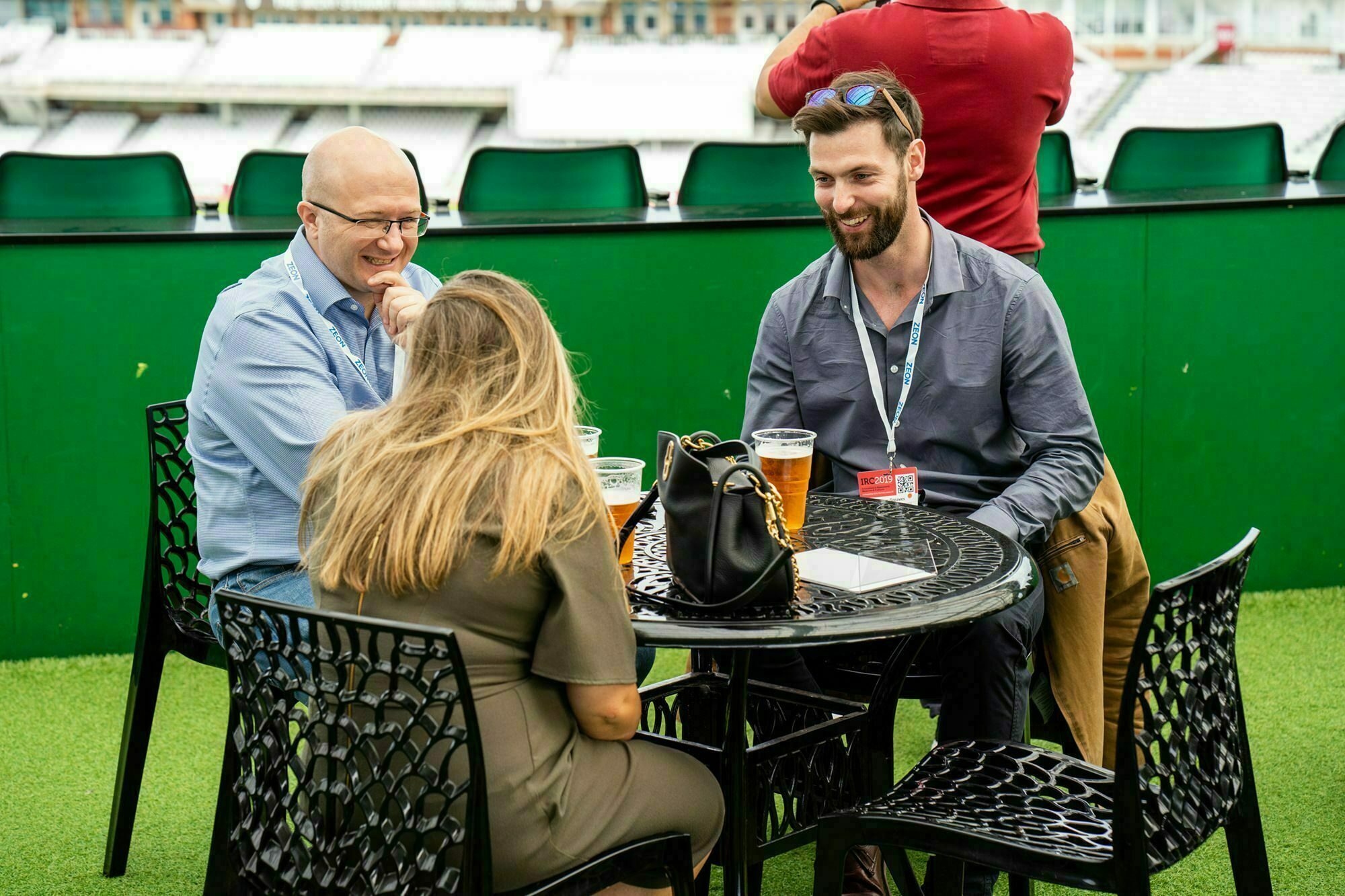 Three people sit around a small black table on a green terrace. Two men and one woman engage in conversation, smiling. Two pints of beer and a handbag are on the table, and an empty grandstand is visible in the background. James Gifford-Mead Photography - Event Photographer London