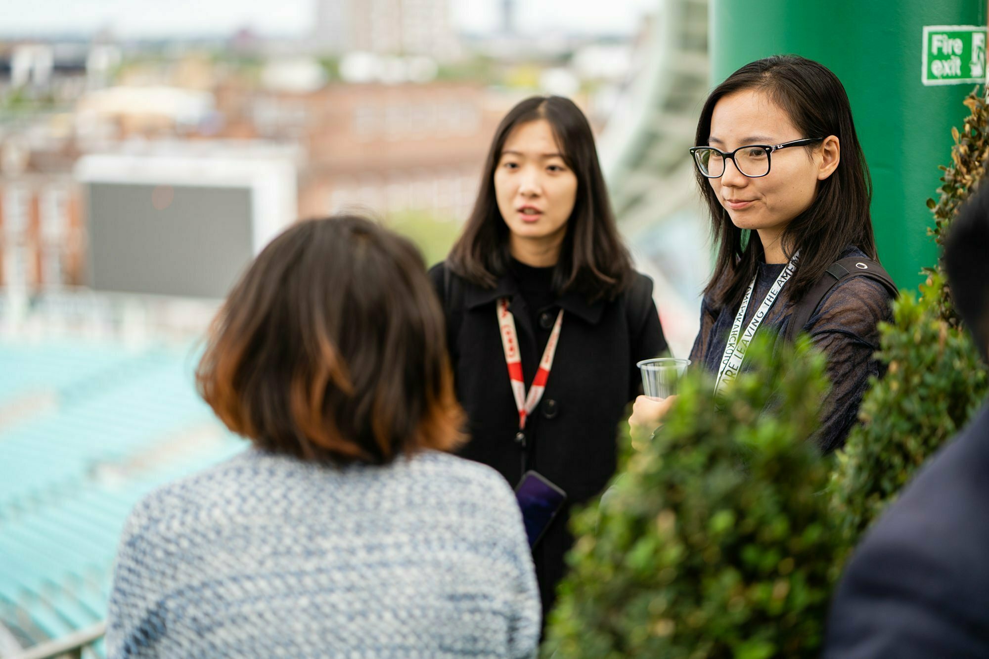 Three people are having a conversation outdoors on a balcony. Two are facing the camera, while the third has their back turned. They are surrounded by greenery and dressed in casual attire. A blurred cityscape is visible in the background. James Gifford-Mead Photography - Event Photographer London