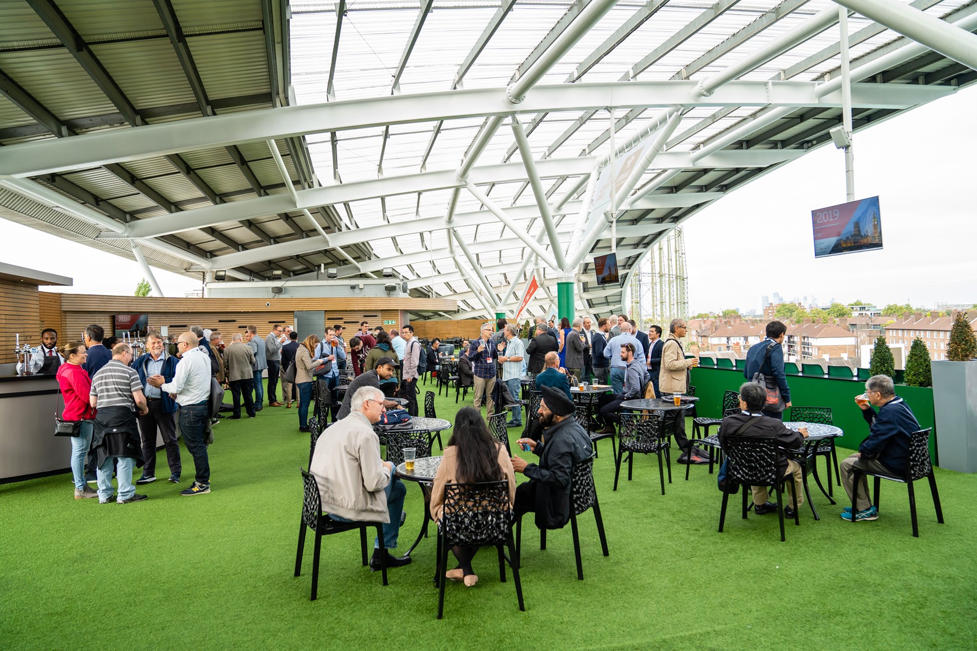 People socializing at an outdoor rooftop venue with a green turf floor and modern overhead structure. Tables are scattered with drinks and snacks, and a bar is visible to the left. The setting offers a city view in the background. James Gifford-Mead Photography - Event Photographer London